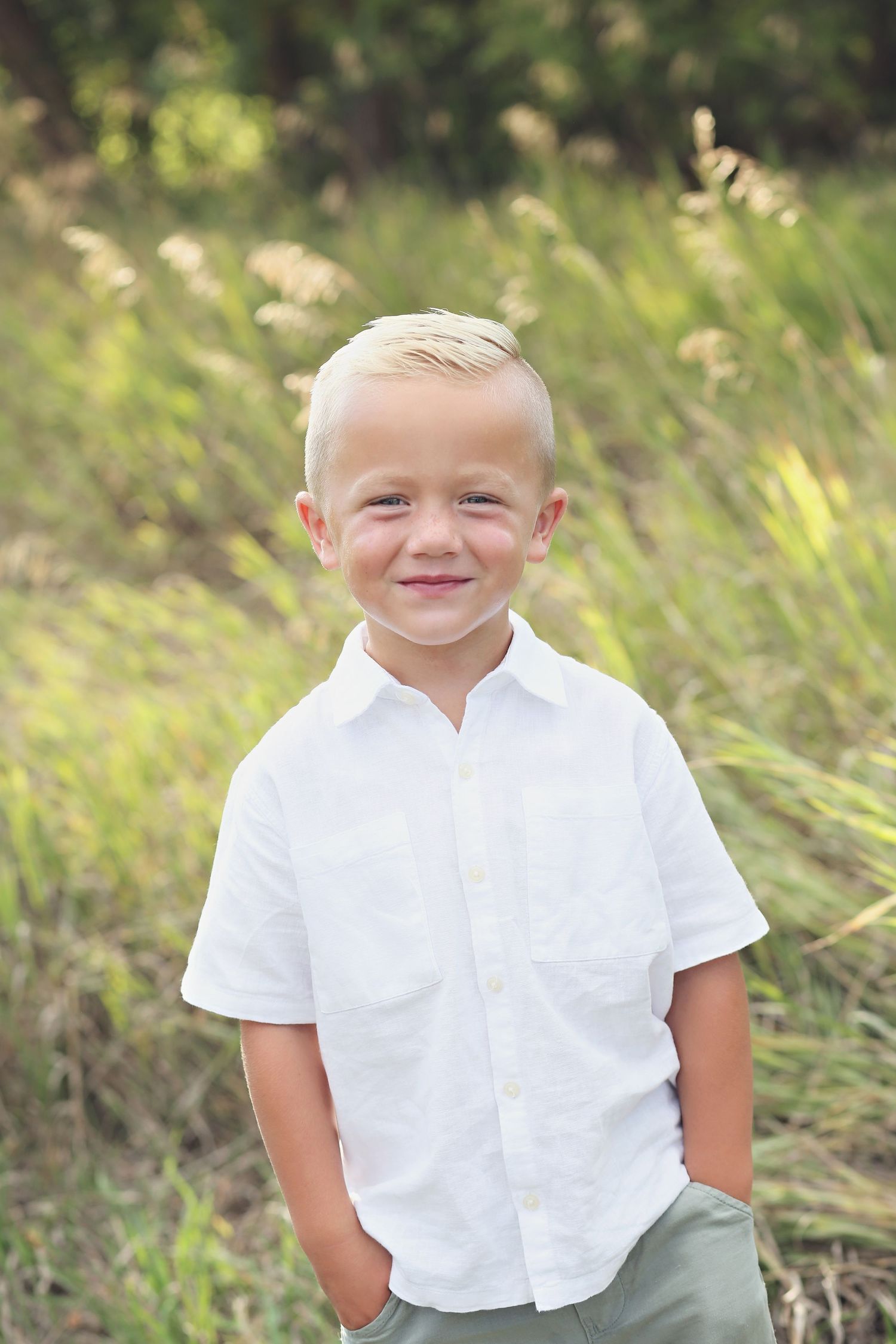 A young child in a white button-down shirt poses outdoors against a natural blurred background.