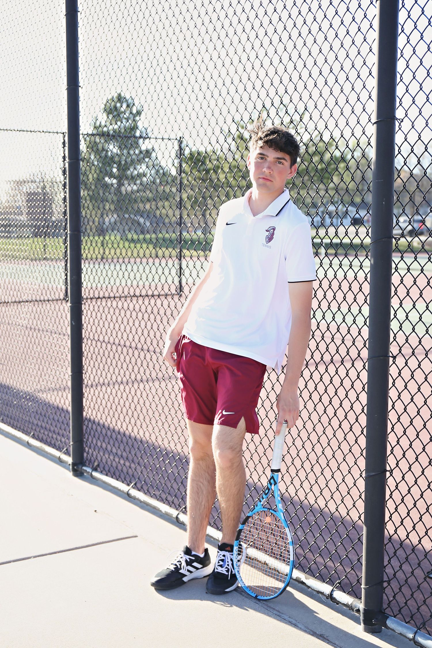 Tennis player in white shirt and maroon shorts practices on outdoor court.