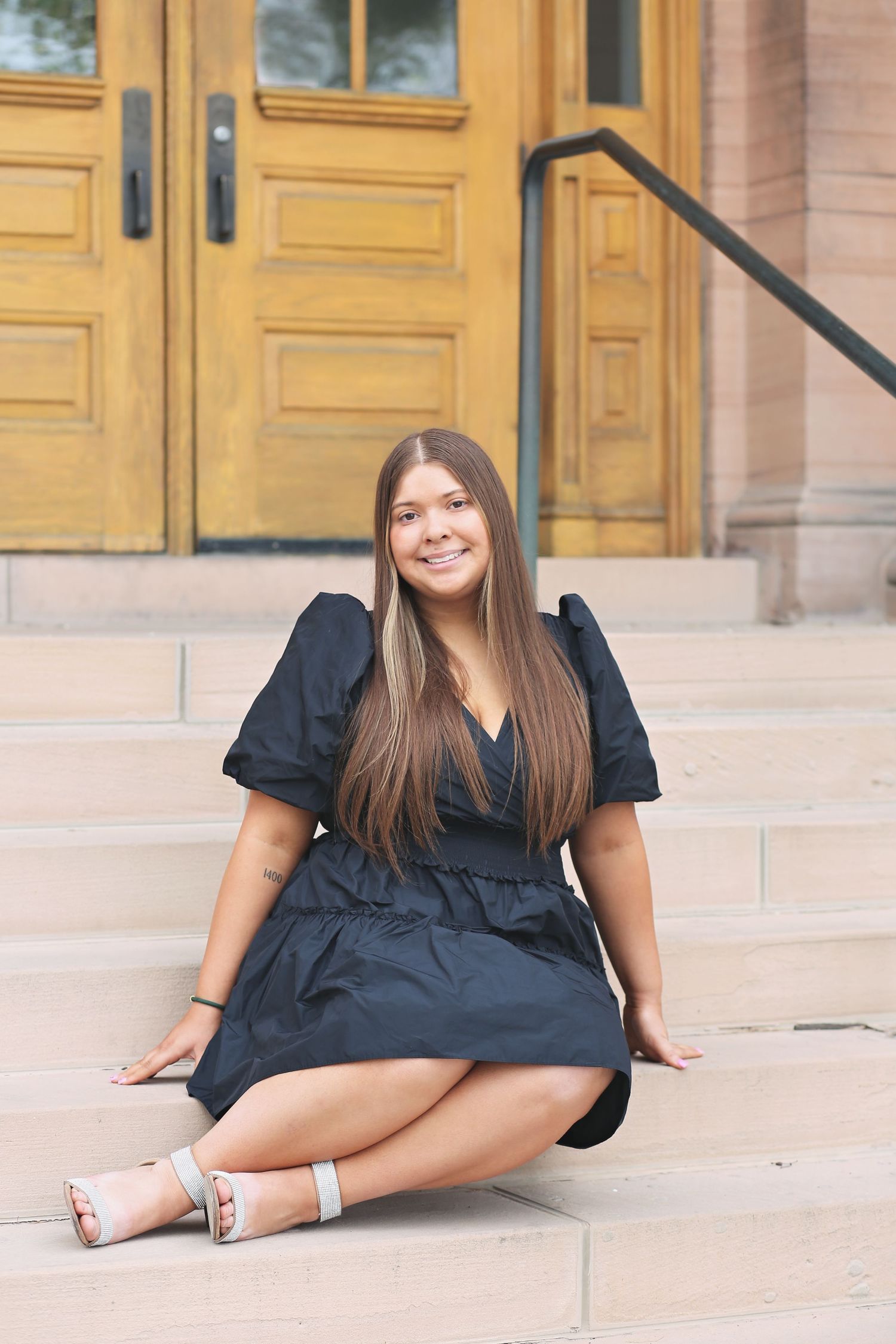 Graduate posing in a black dress on stone steps in front of yellow doors.