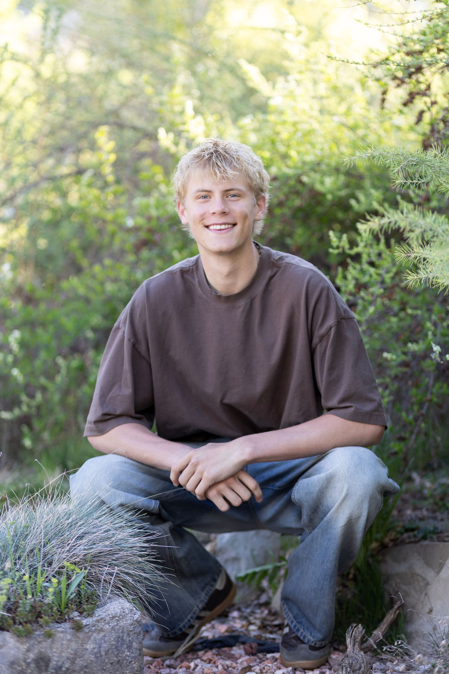 Smiling young blond man sitting cross-legged on rocks outdoors, wearing brown t-shirt and jeans, surrounded by green foliage.