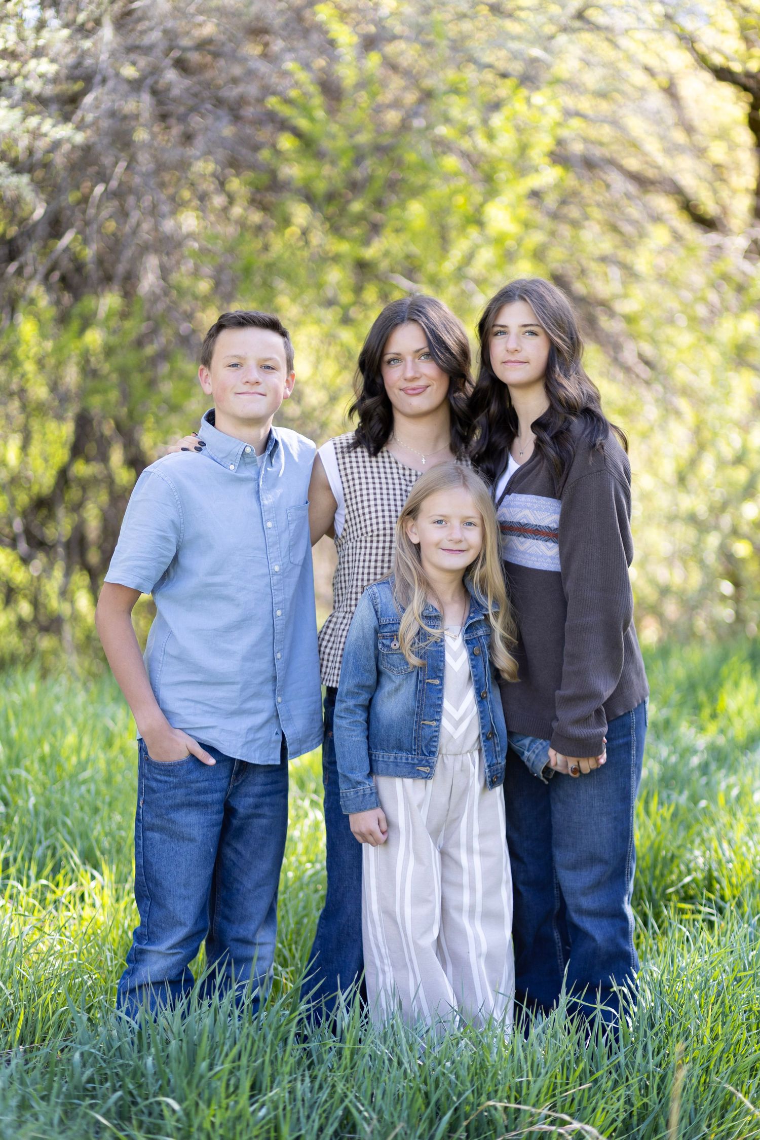 Four siblings pose together in a lush green outdoor setting, smiling for a family portrait photo.
