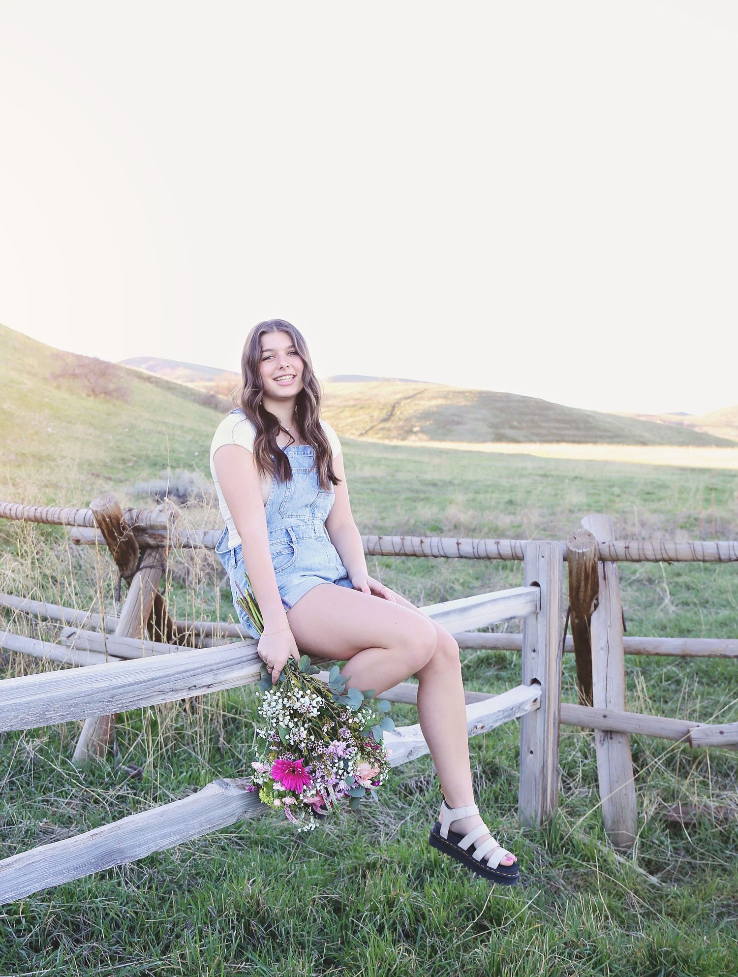Person in summer outfit sits on wooden fence in sunny countryside setting.