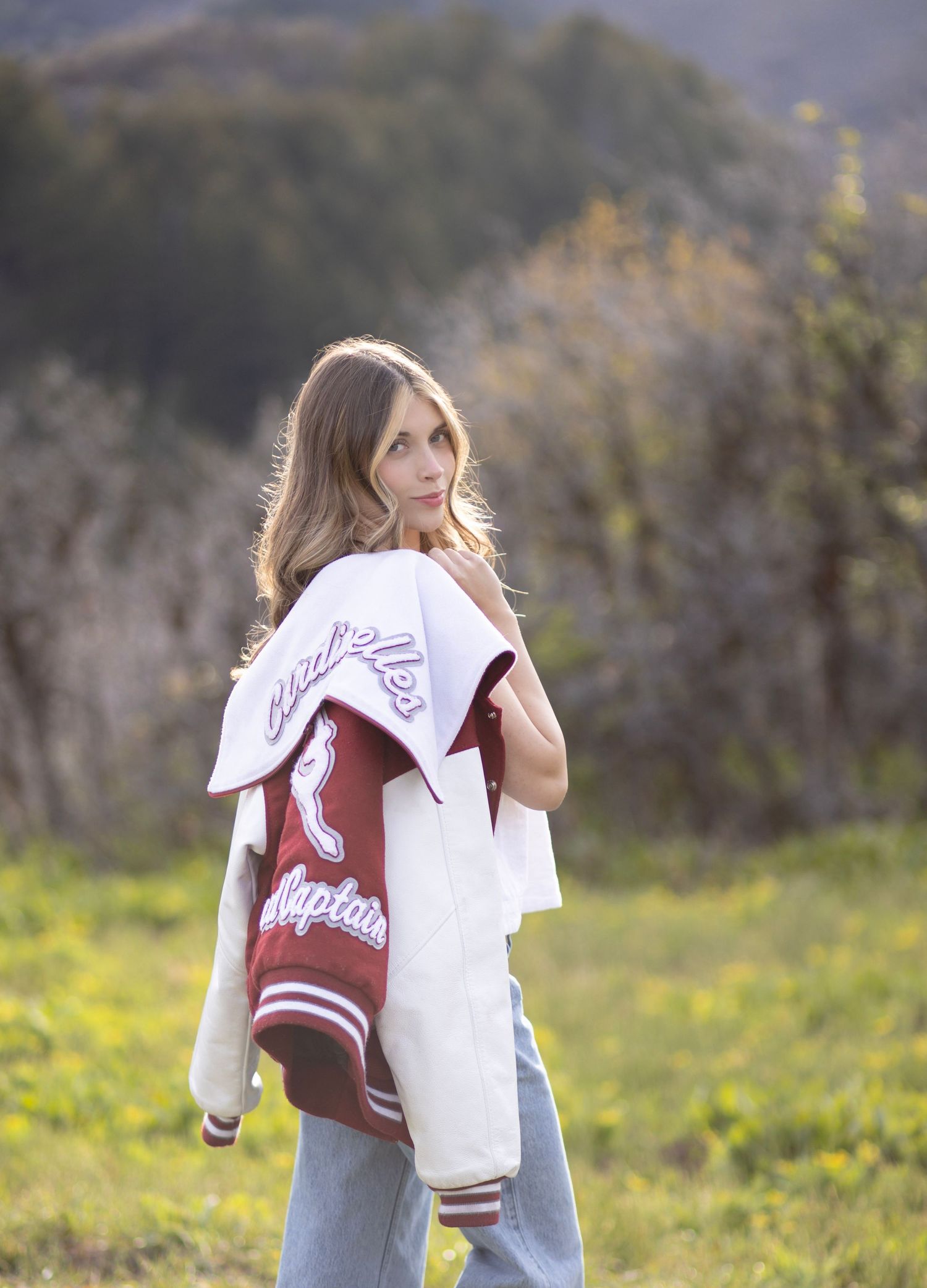 Woman posing outdoors in jeans and a red varsity jacket draped over her shoulders, with green hills in the background.