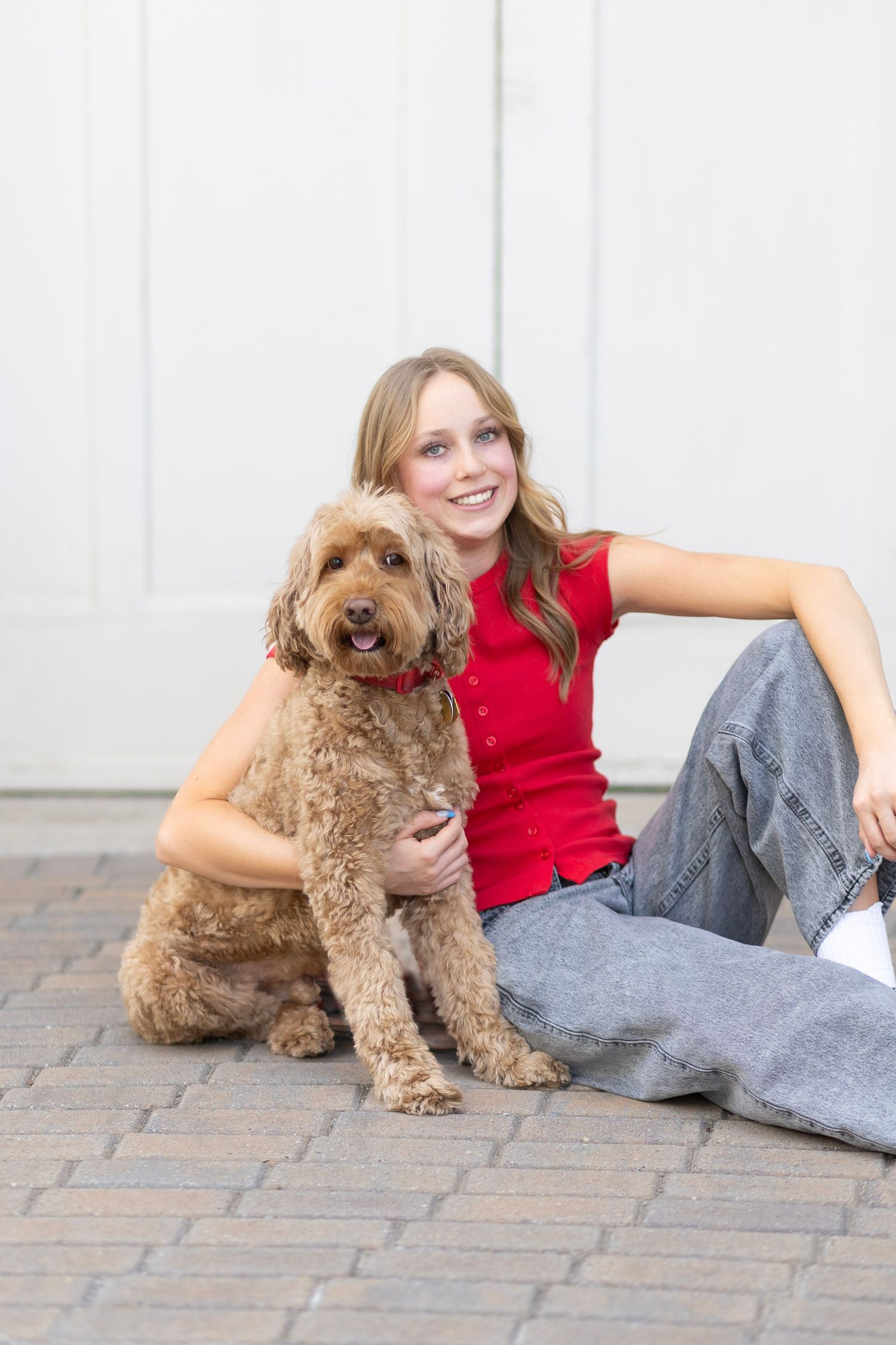 Smiling young woman in red top and jeans sitting on brick pavers with a fluffy golden doodle dog.