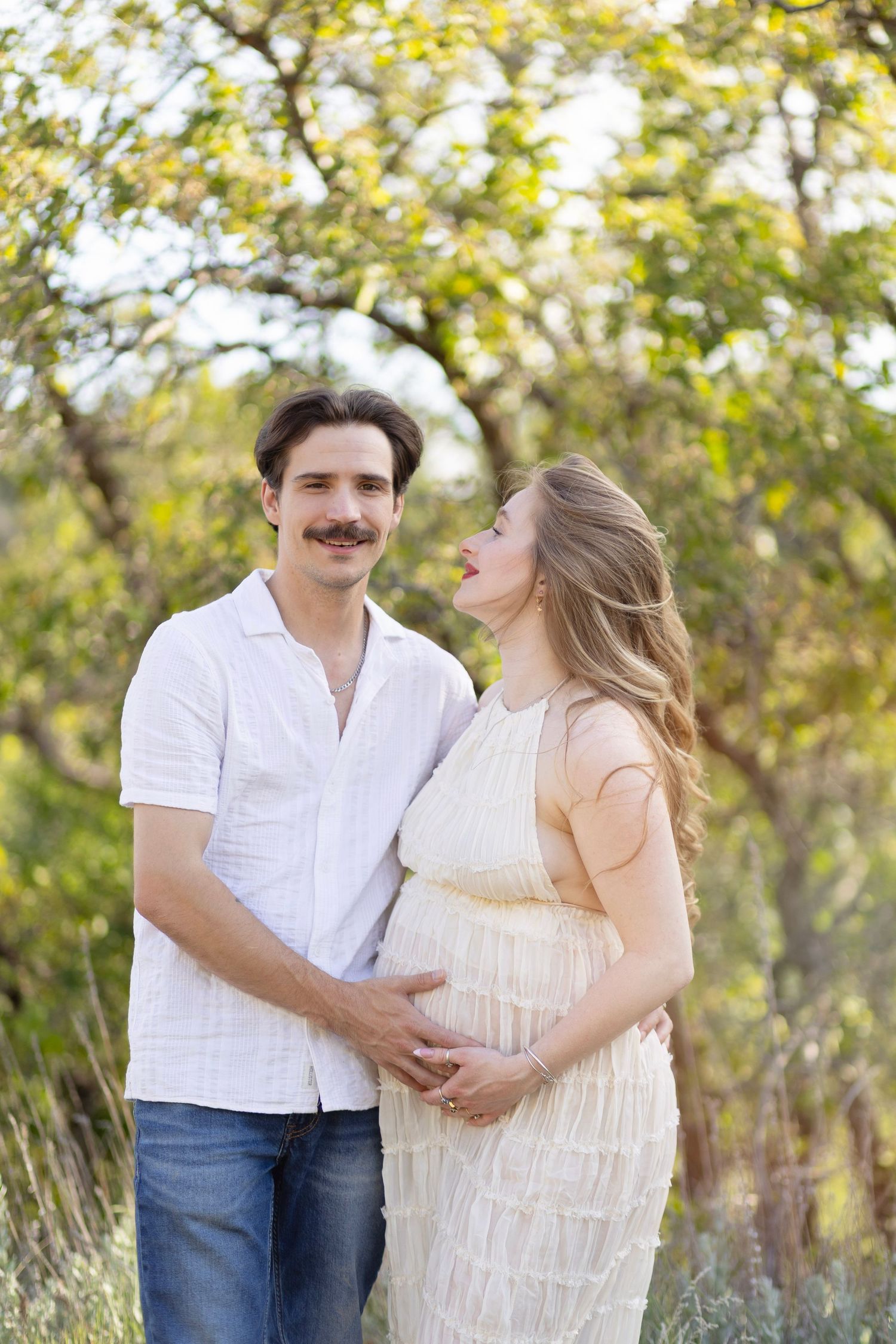 Couple outdoors, pregnant woman in cream dress, man in white shirt and jeans, embracing in a sunny, tree-lined setting.