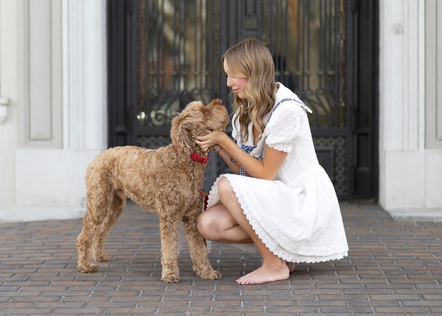 Young woman in white dress crouching to pet her fluffy golden doodle dog with red collar outdoors.
