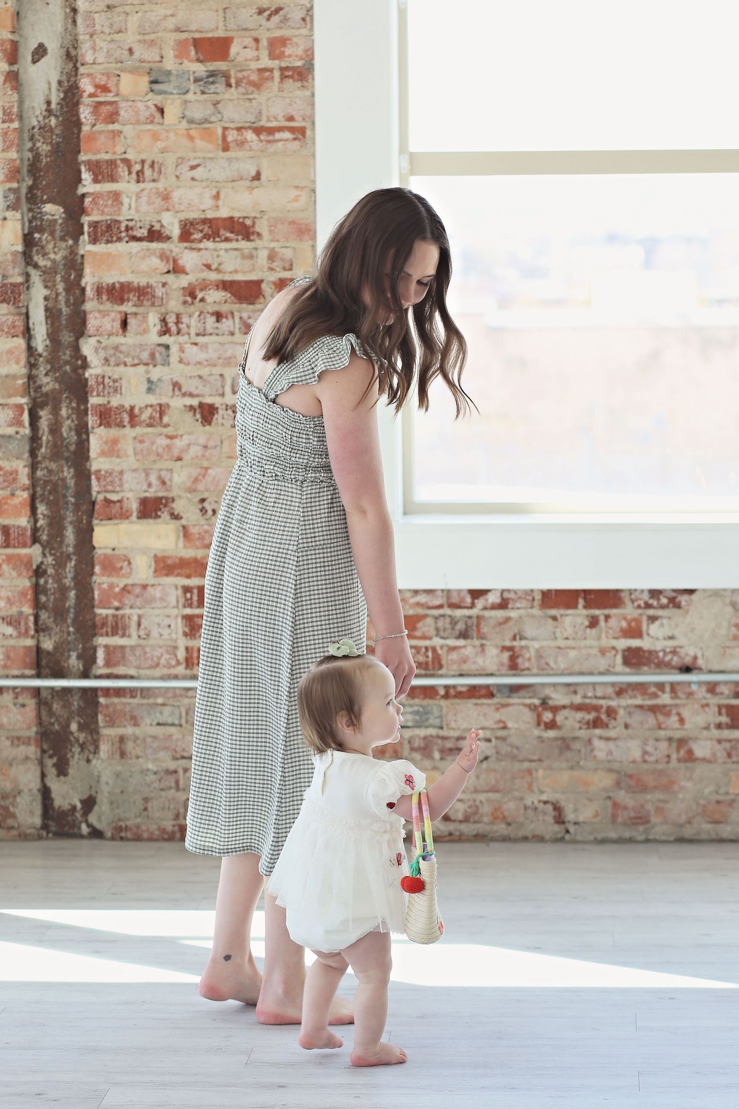 A parent and toddler standing together in a sunlit room with exposed brick walls.