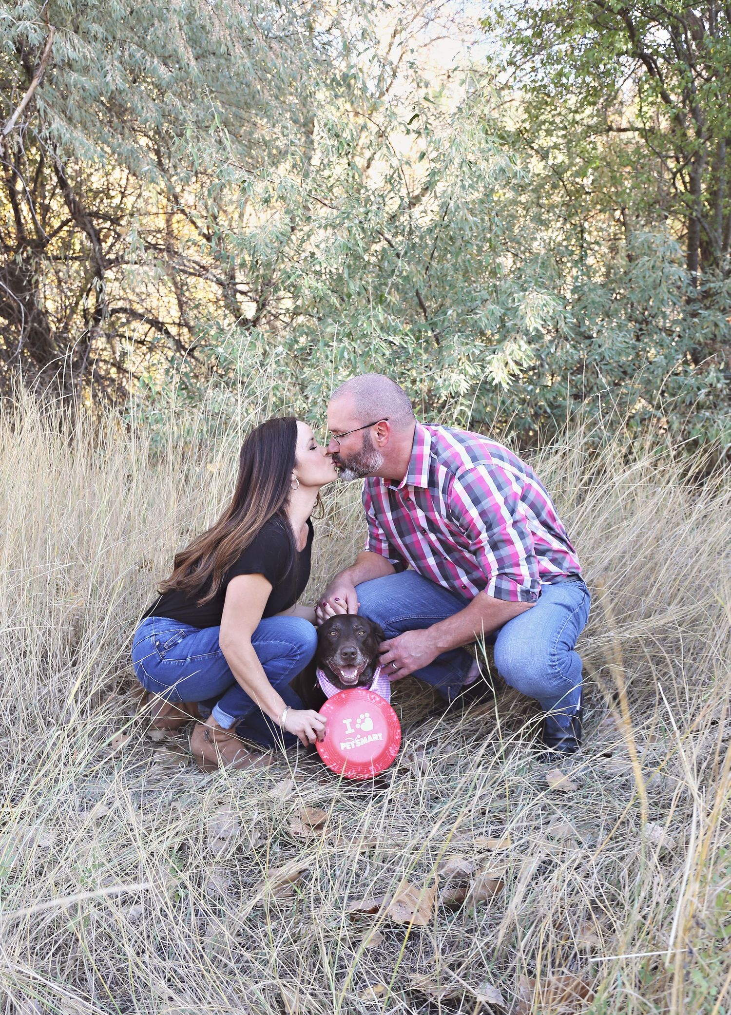 Family plays with chocolate lab and frisbee in tall grass field.