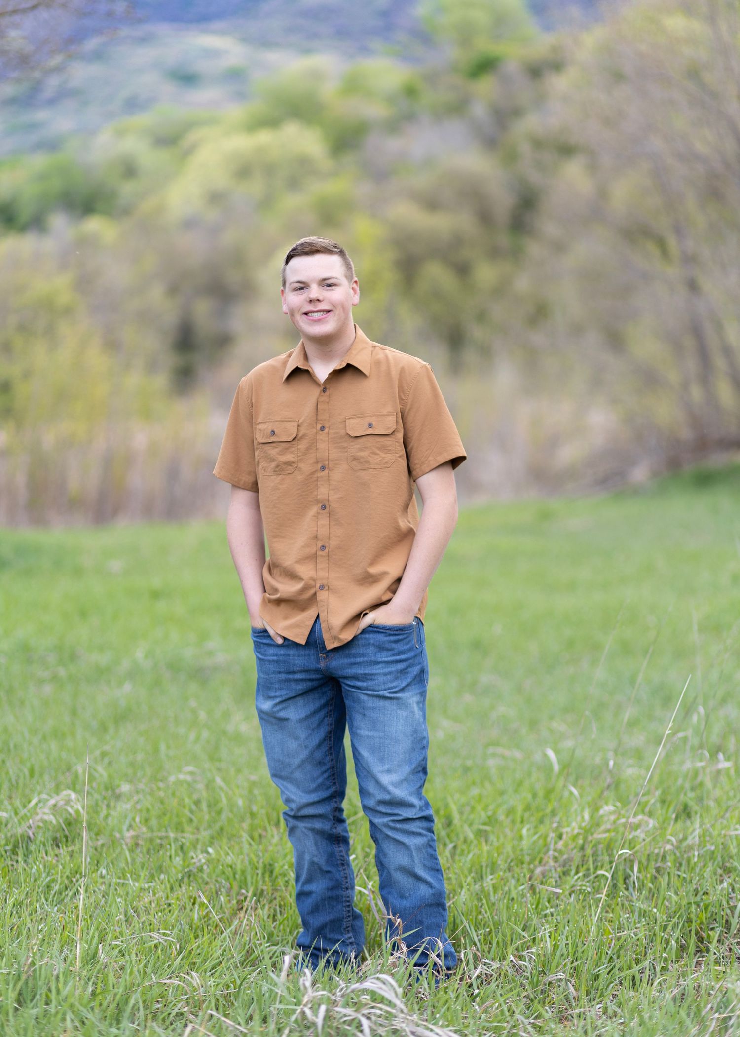 Young man in brown shirt and jeans smiling outdoors in a green field with trees and hills in background.