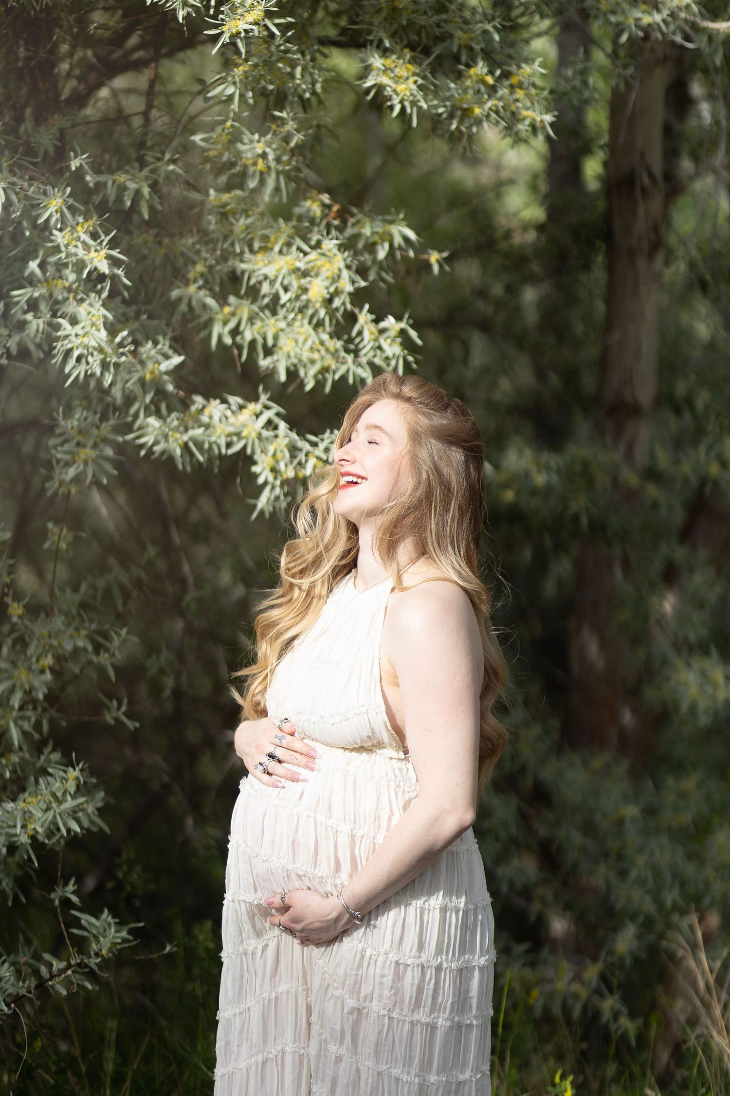 Pregnant woman in white dress holding baby bump outdoors among green trees, smiling joyfully in natural sunlight.