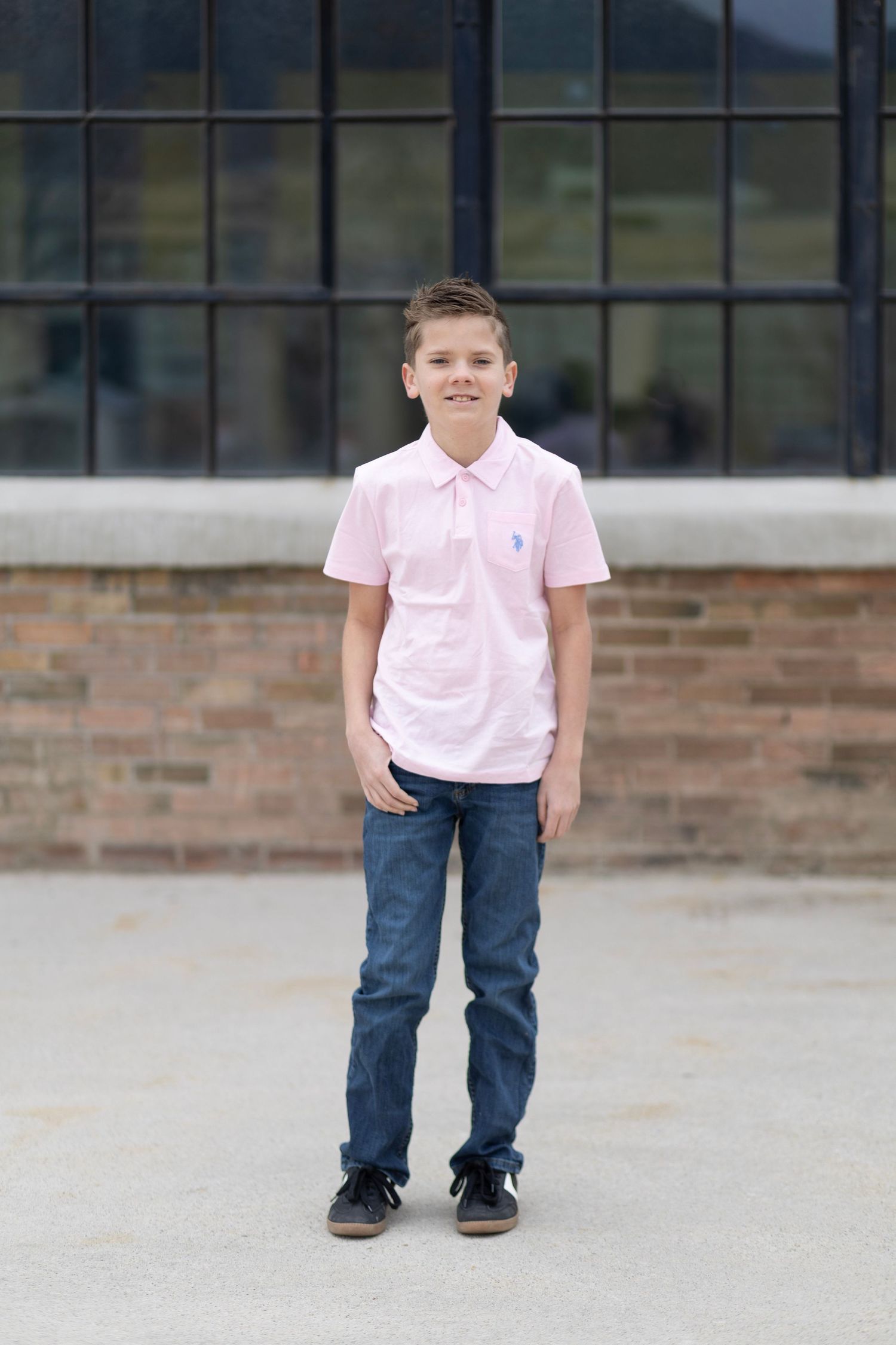 A smiling boy in a pink polo shirt and jeans stands confidently outside a brick building.