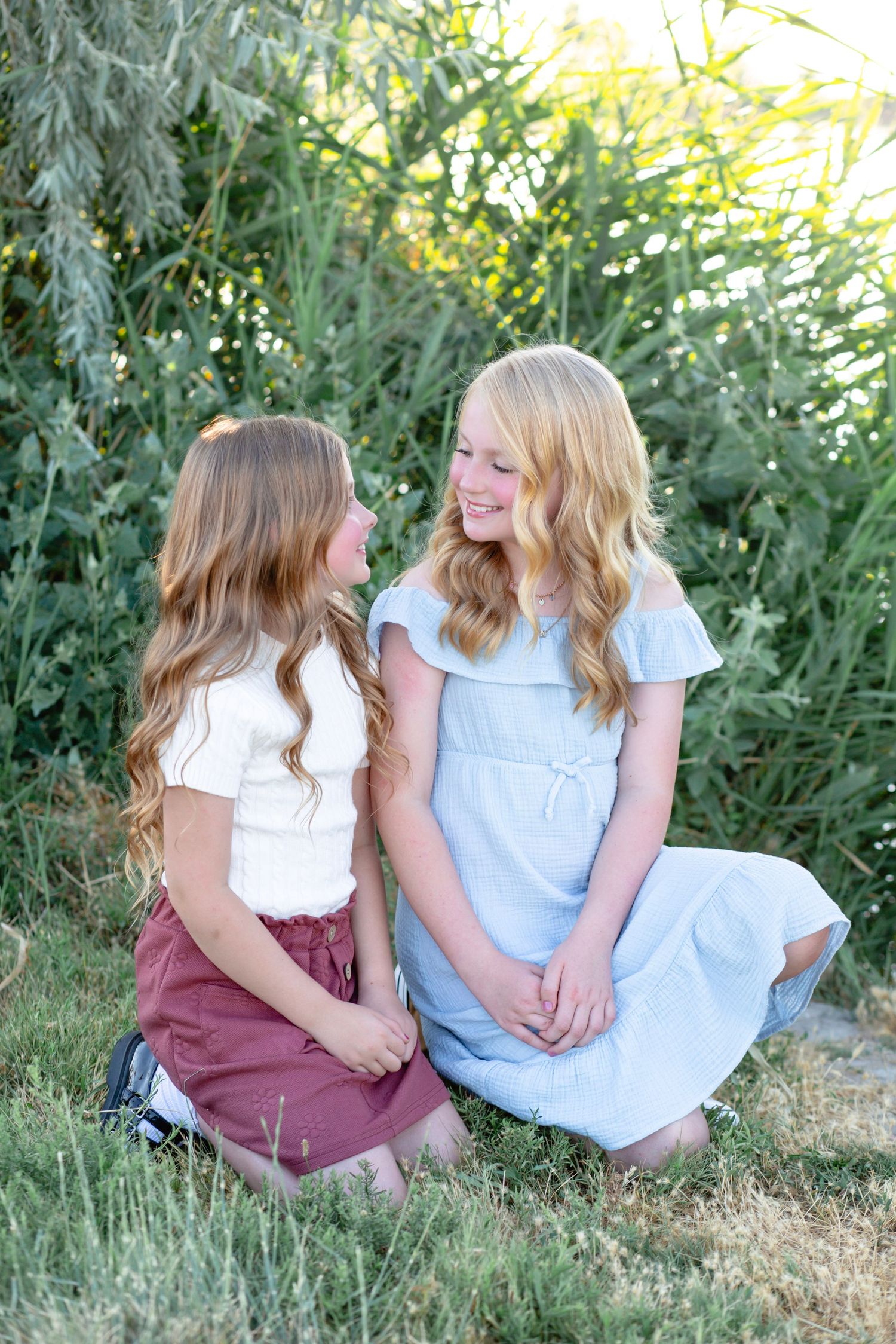 Two young people sit together on grass having a conversation in a natural outdoor setting.