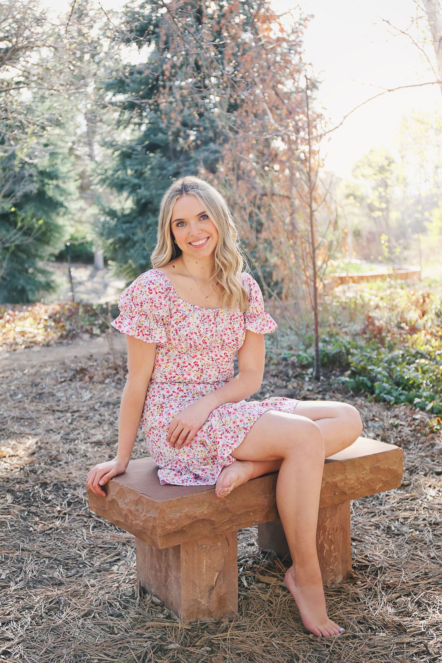 A person in a floral dress sits on a rustic wooden bench in a sunlit outdoor setting with trees in the background.