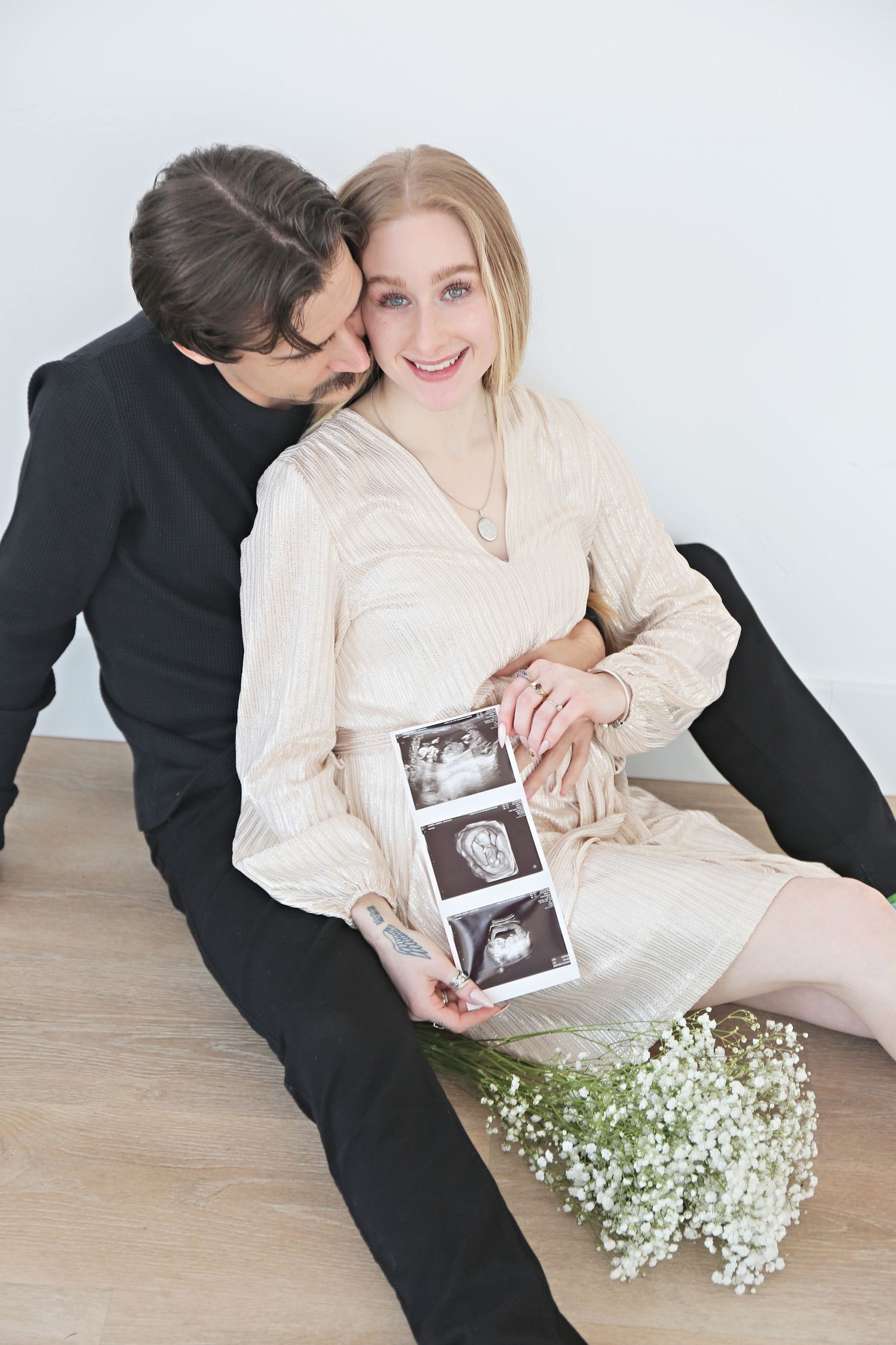 Couple sharing intimate moment while holding ultrasound photos with baby's breath flowers nearby.