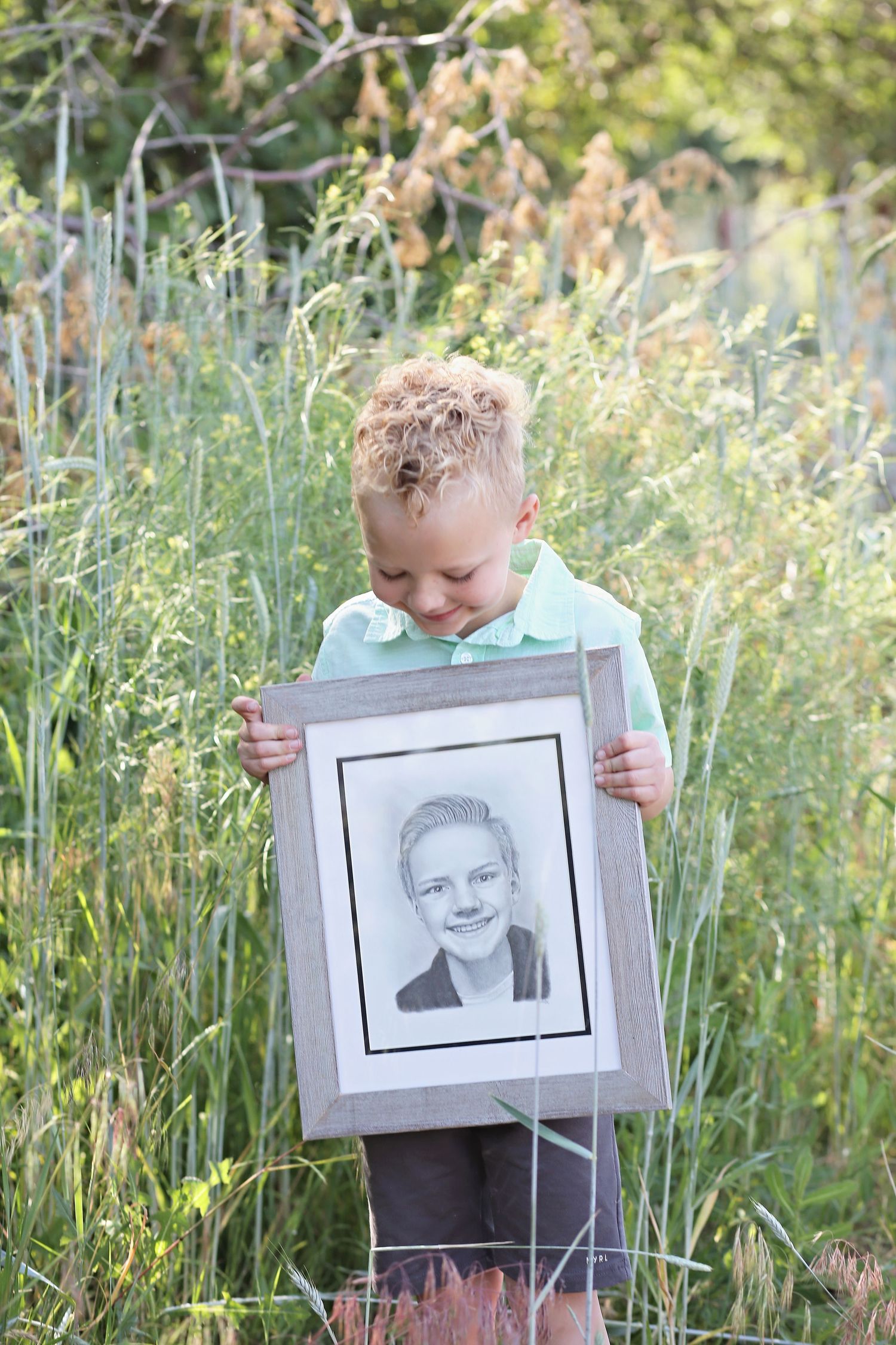 A young child holds a framed portrait while standing in tall grass on a sunny day.