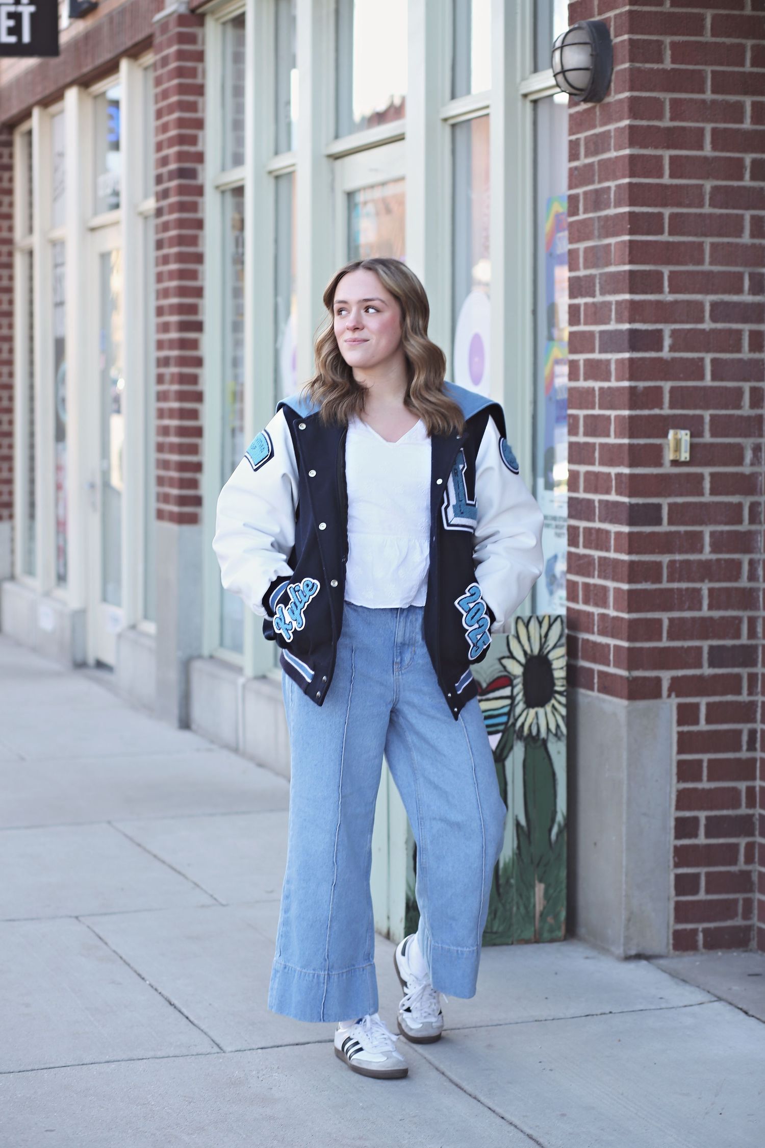 Person wearing varsity jacket and wide-leg jeans standing casually against brick storefront.