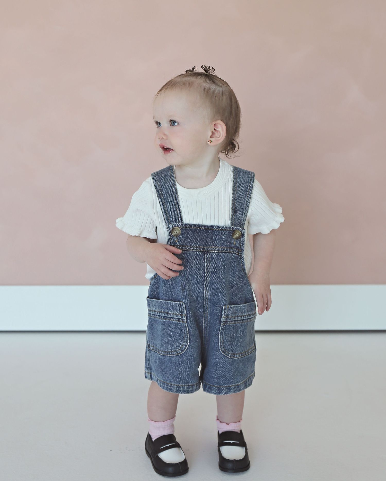 Small child wearing denim overalls and white shoes stands against a pink wall.