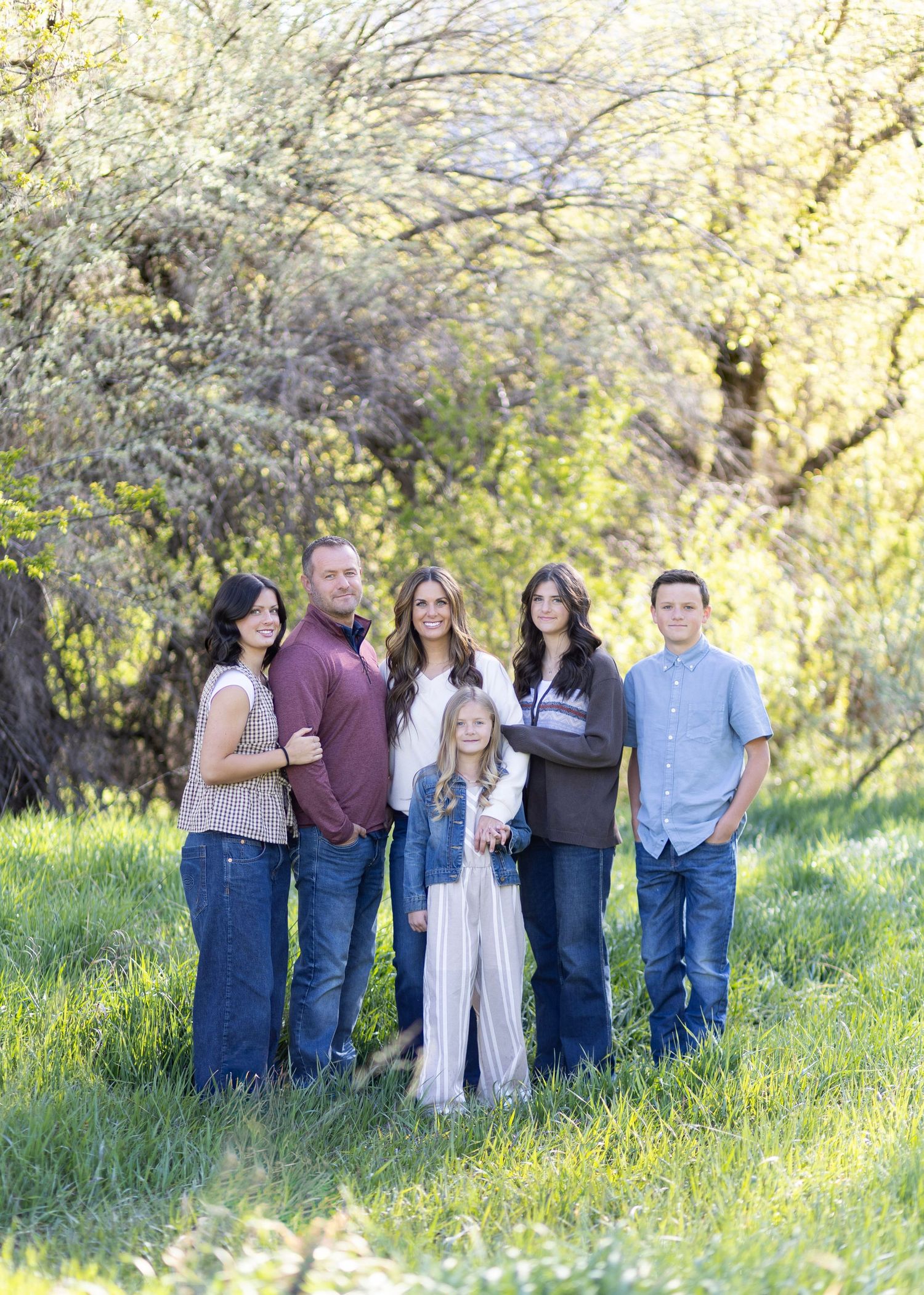 Family of six posing outdoors in a lush green meadow with blooming trees in spring sunlight.