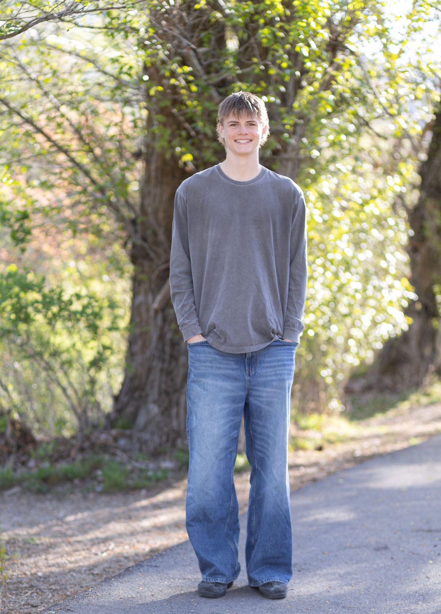 Smiling young man in gray long-sleeve shirt and flared jeans standing on a tree-lined path outdoors.