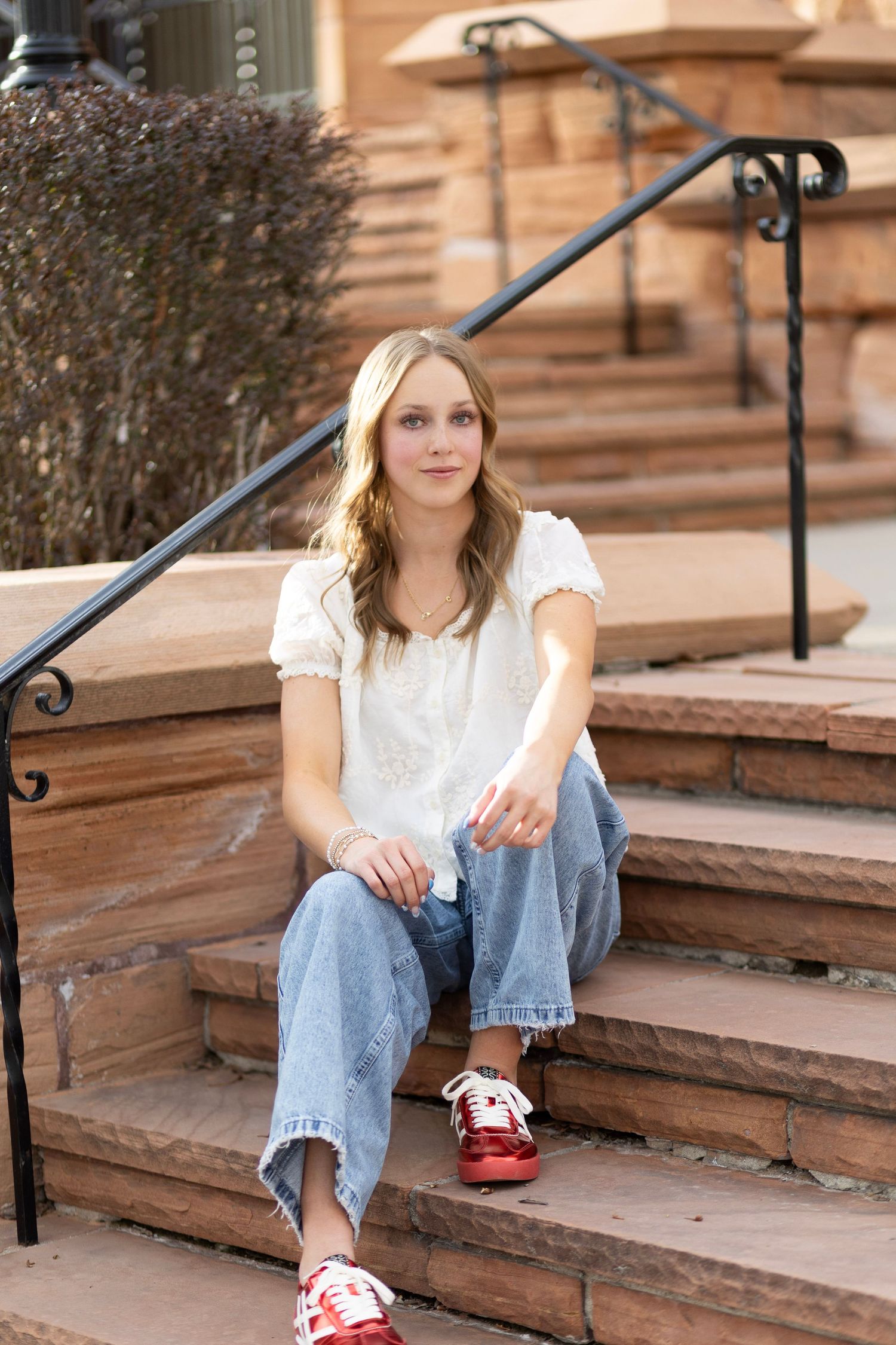 Young blonde woman in white blouse, jeans, and red sneakers sits casually on stone steps outdoors.