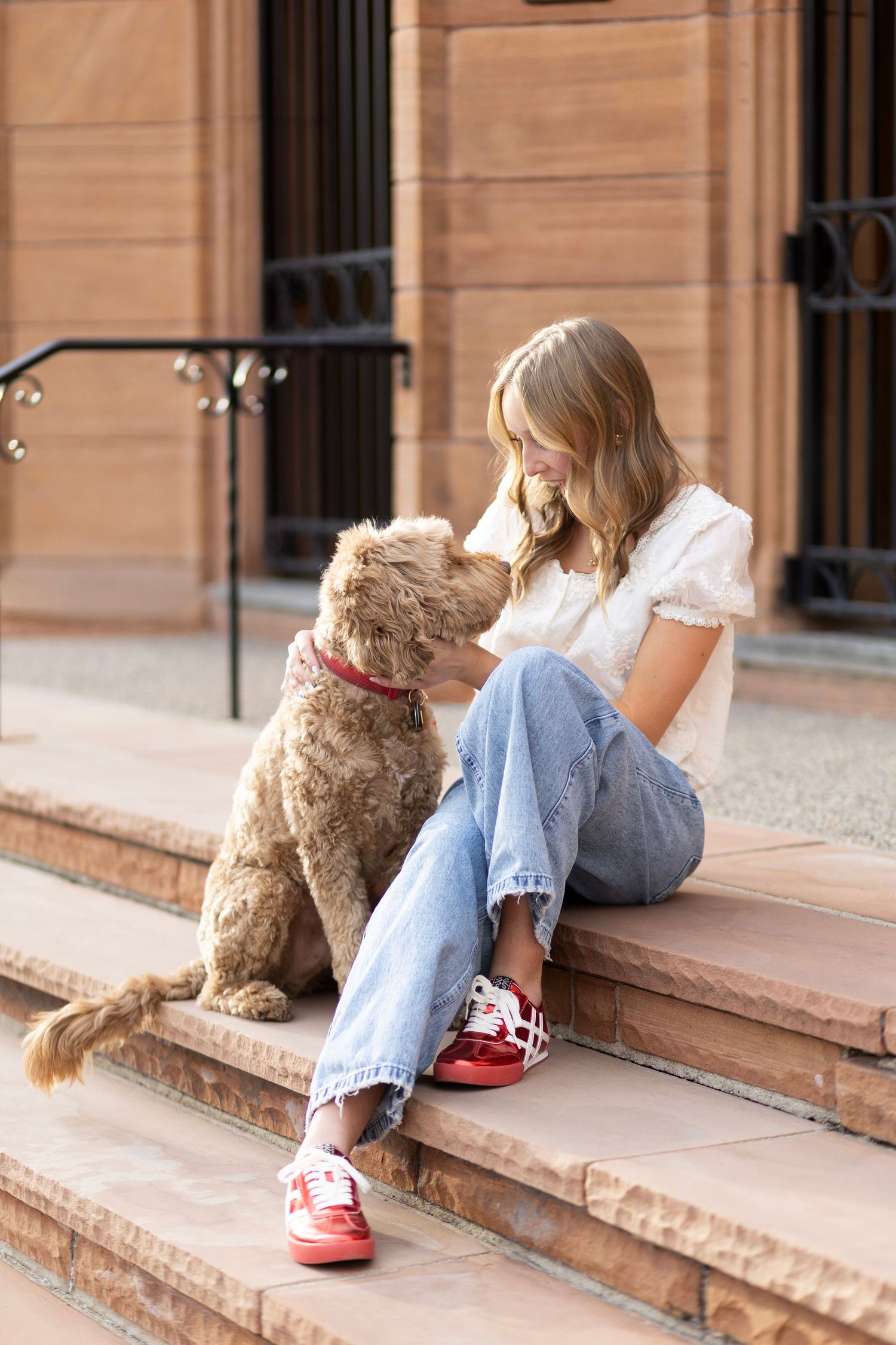 Young woman in jeans and red sneakers sitting on brownstone steps with a fluffy brown goldendoodle puppy.