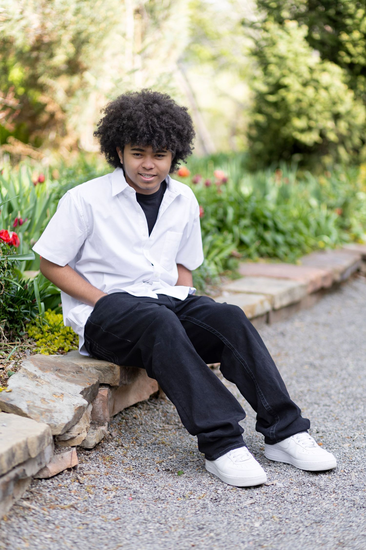 A young African American teen boy with curly hair sits on a stone wall in a garden, wearing a white shirt and black jeans.