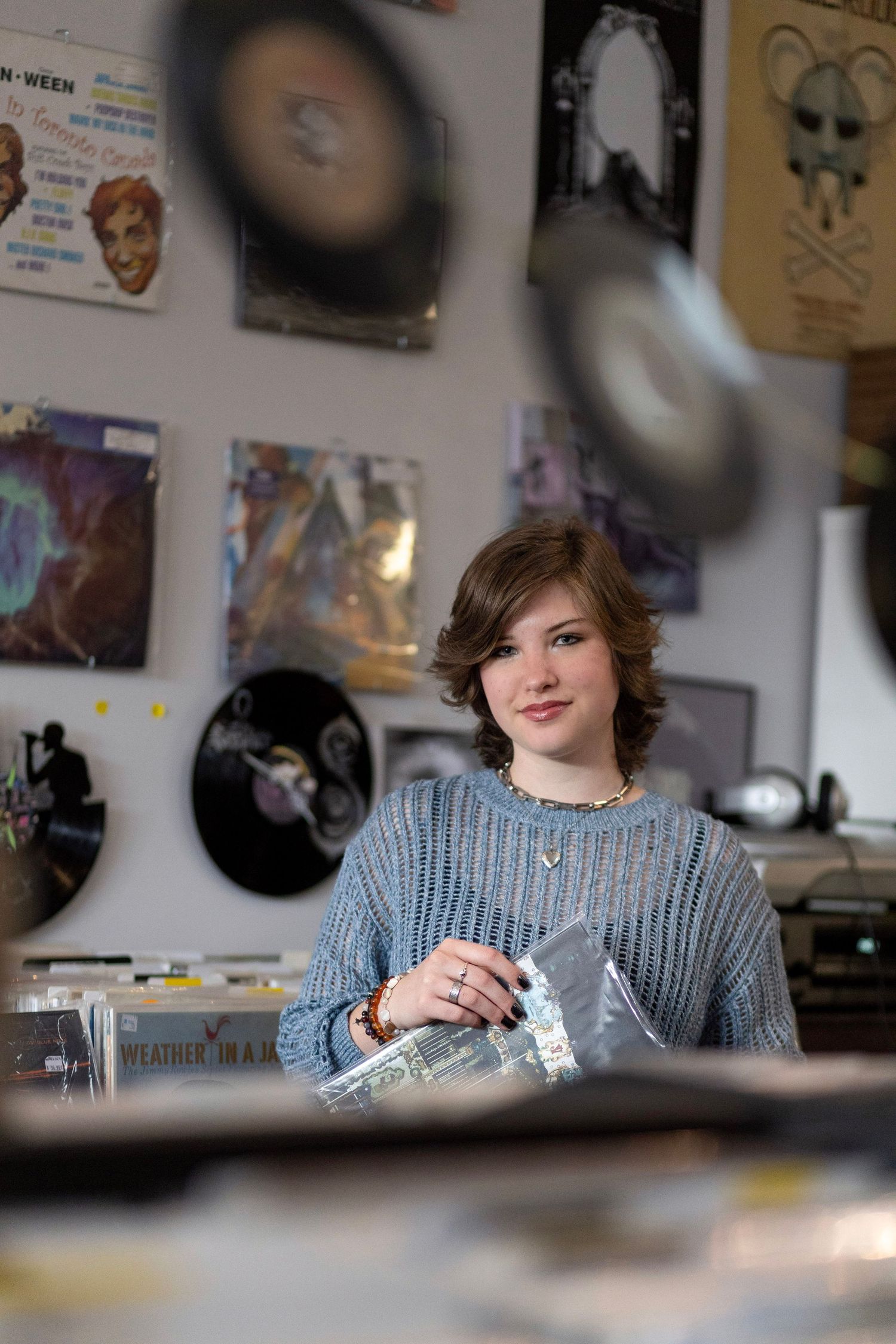 Young person in blue sweater browsing vinyl records in a music shop surrounded by album covers on the wall.