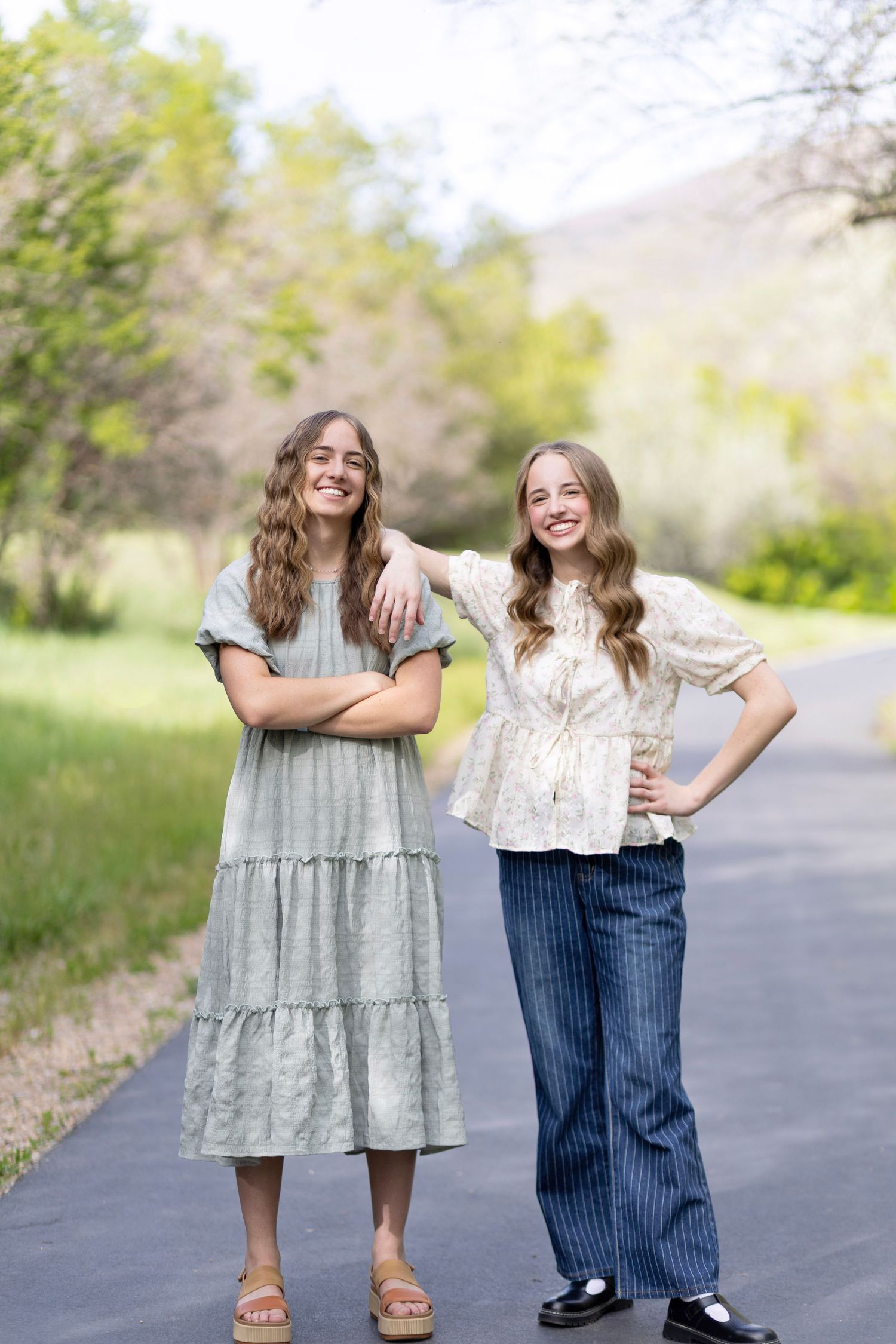 Two smiling young women with long hair posing outdoors on a path, one in a sage dress and one in a floral top with blue pants.