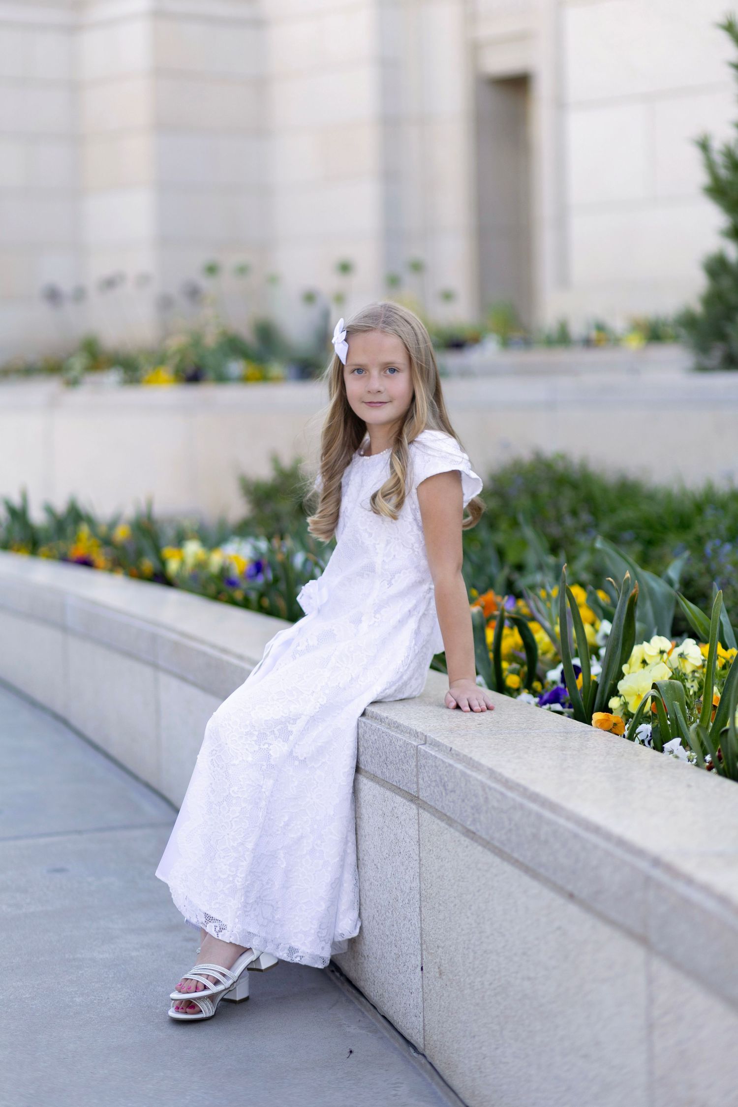 Young girl in white dress sitting on stone ledge surrounded by colorful flowers outside a building.