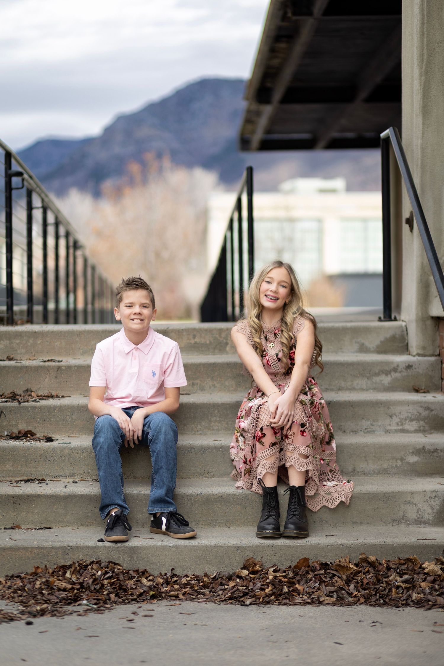 Two smiling children sit on outdoor concrete steps with mountain backdrop, boy in pink polo and girl in floral dress.