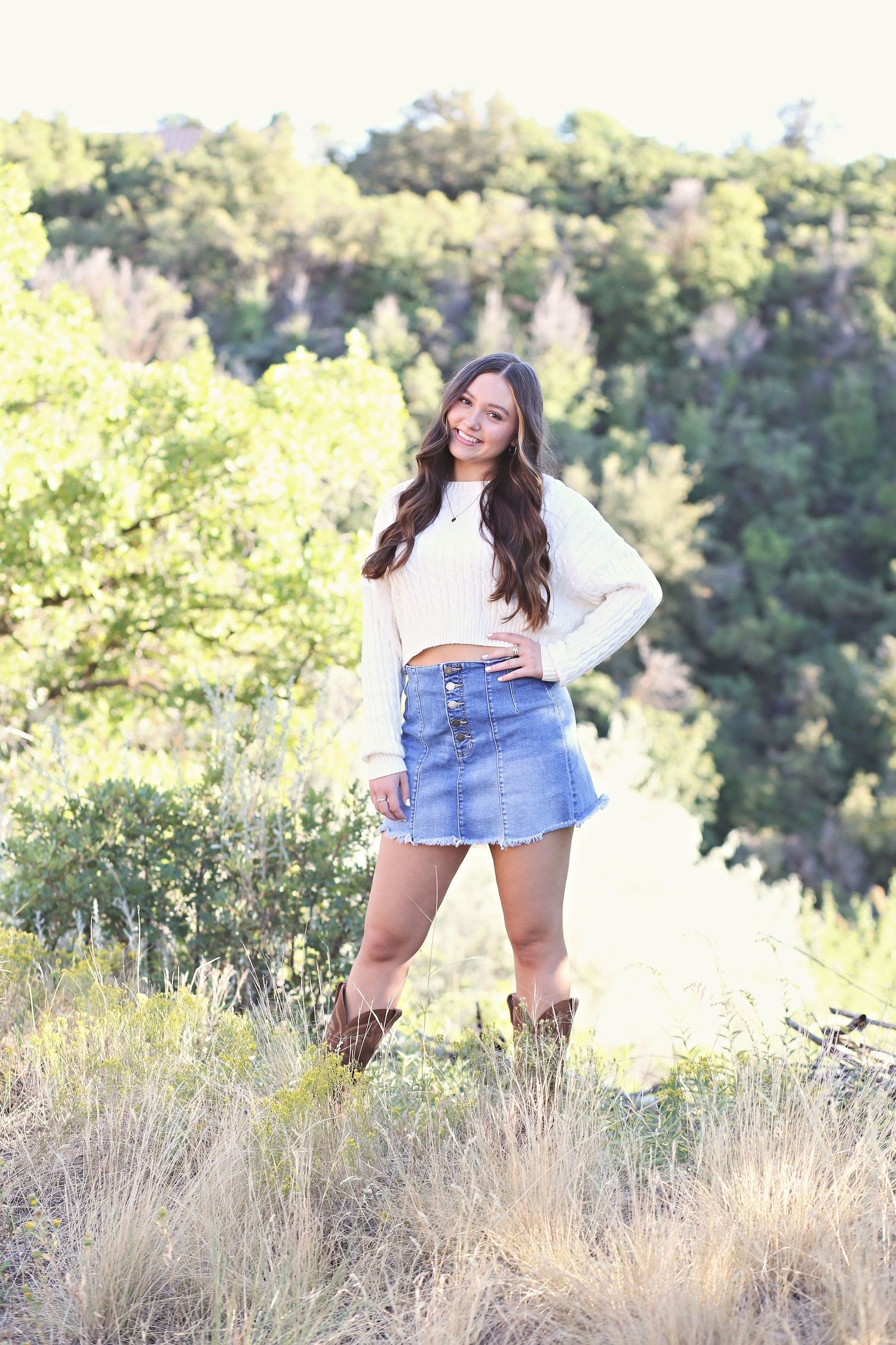 Person in a white sweater and denim skirt poses outdoors on a hillside with brown boots and natural scenery.