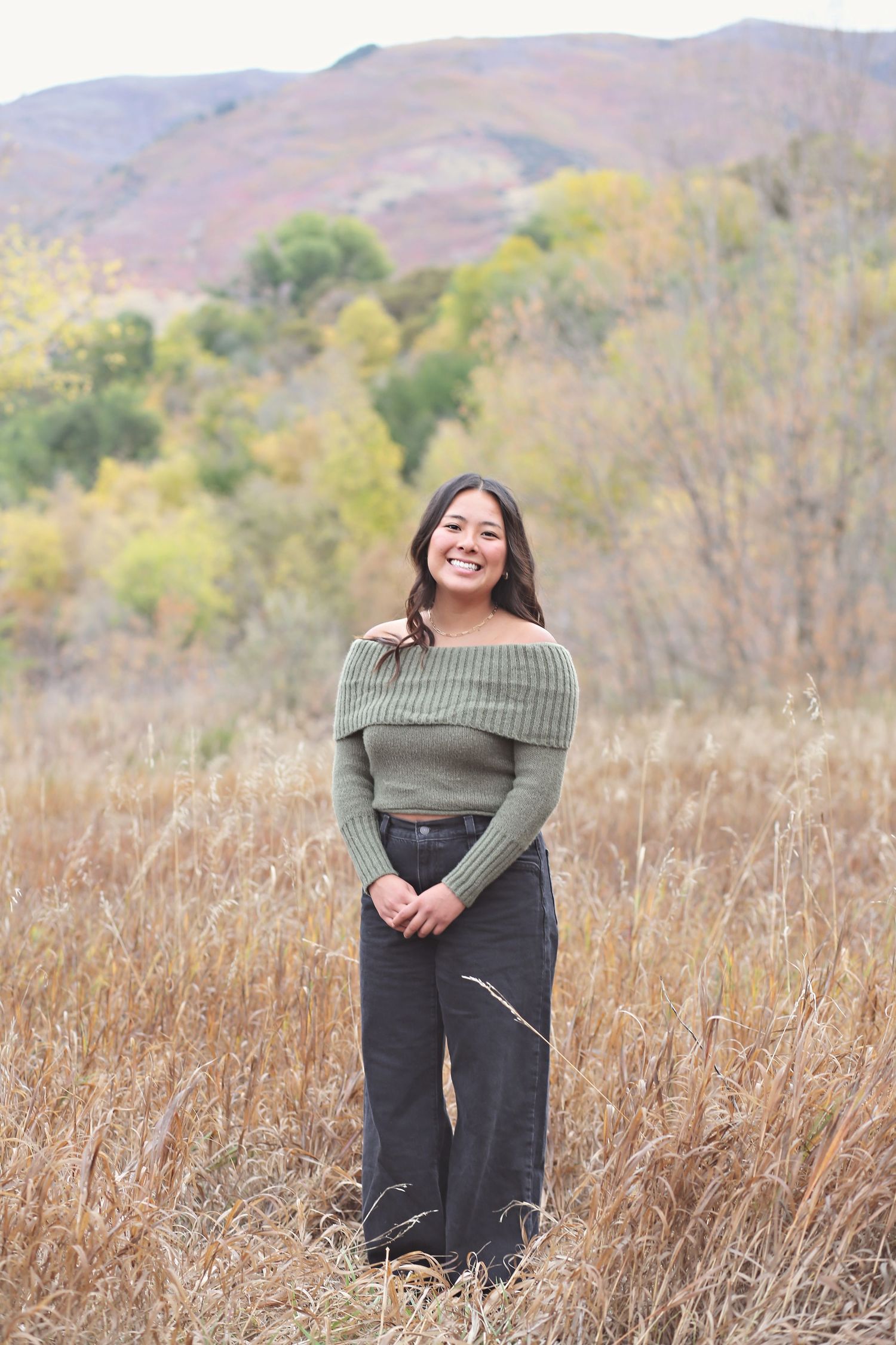 A person in an olive green top and dark pants poses in a field with autumn mountains in the background.