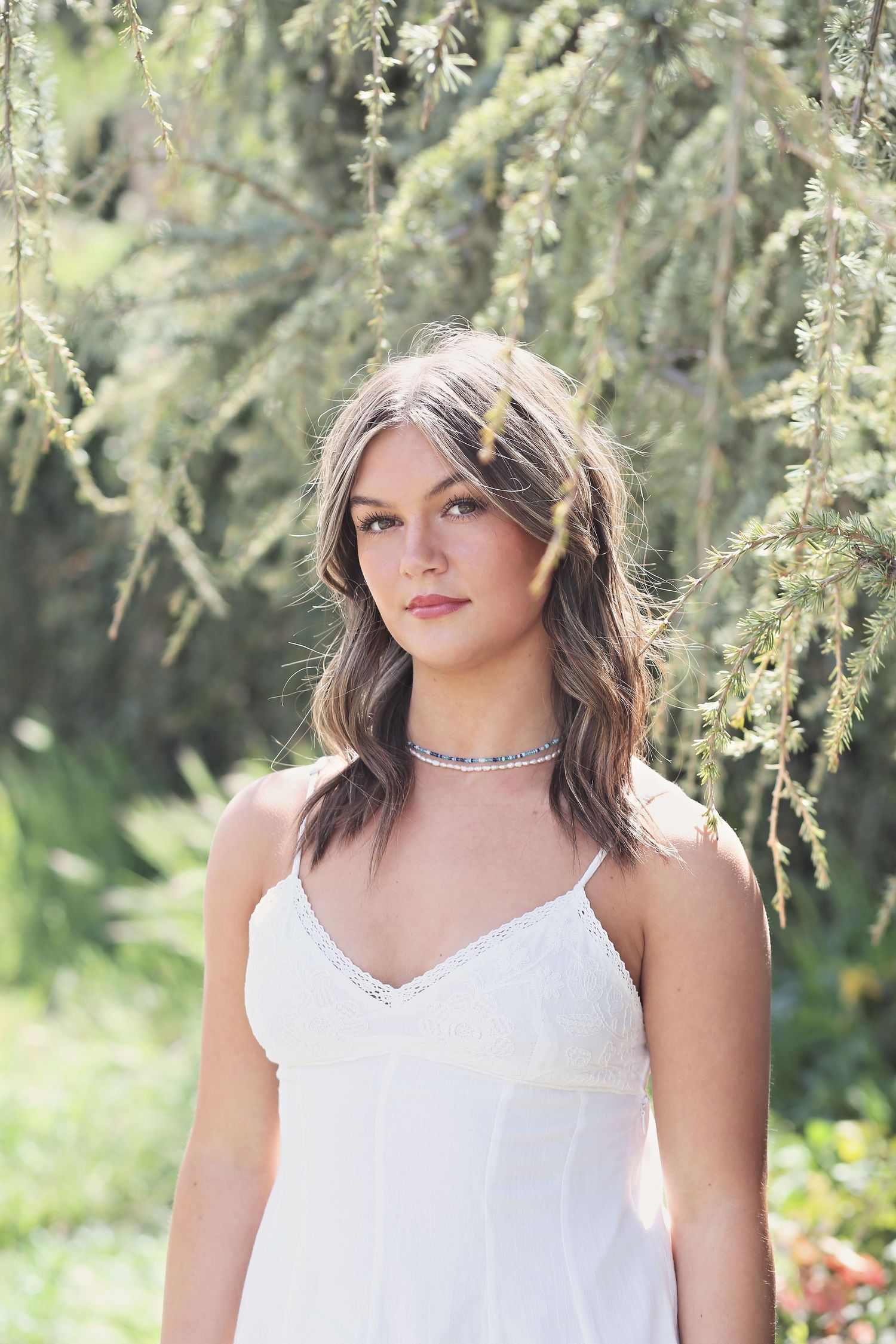 A woman in a white spaghetti strap dress poses outdoors among hanging willow branches on a sunny day.