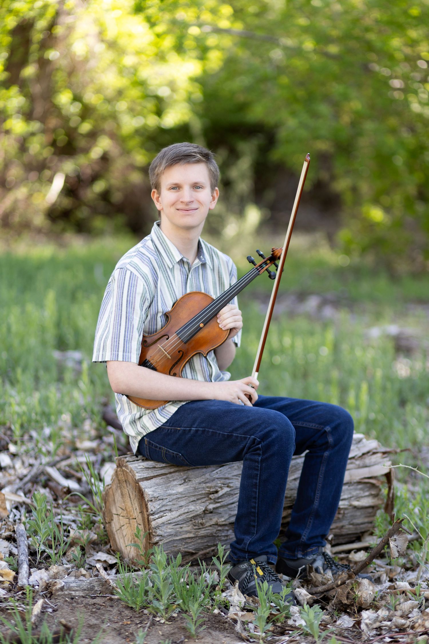 Young man sitting on a log outdoors, smiling while holding a violin and bow, wearing a striped shirt and jeans.