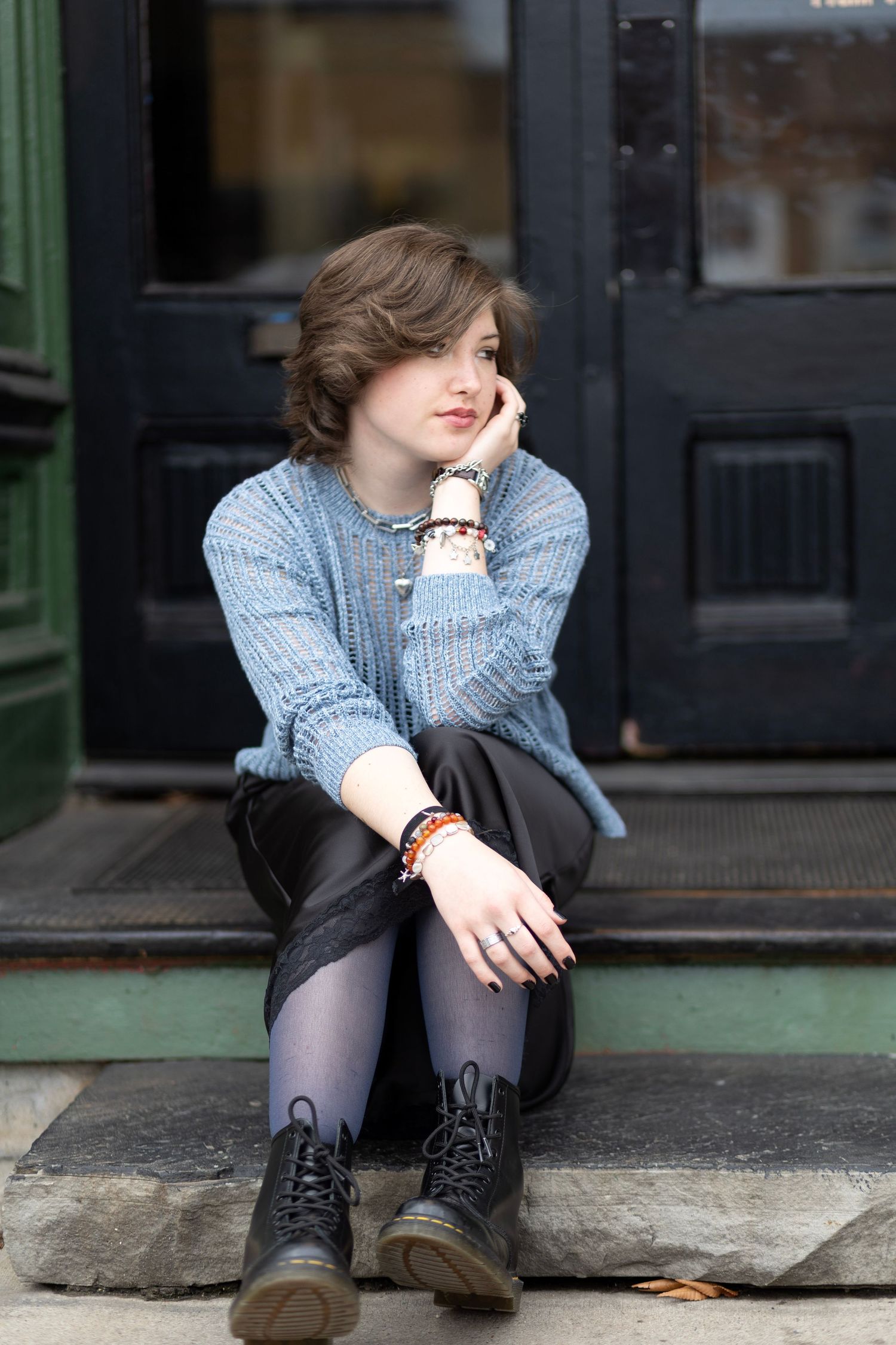 Young woman in blue knit sweater and black skirt sits on stone steps, wearing Doc Martens and colorful bracelets.