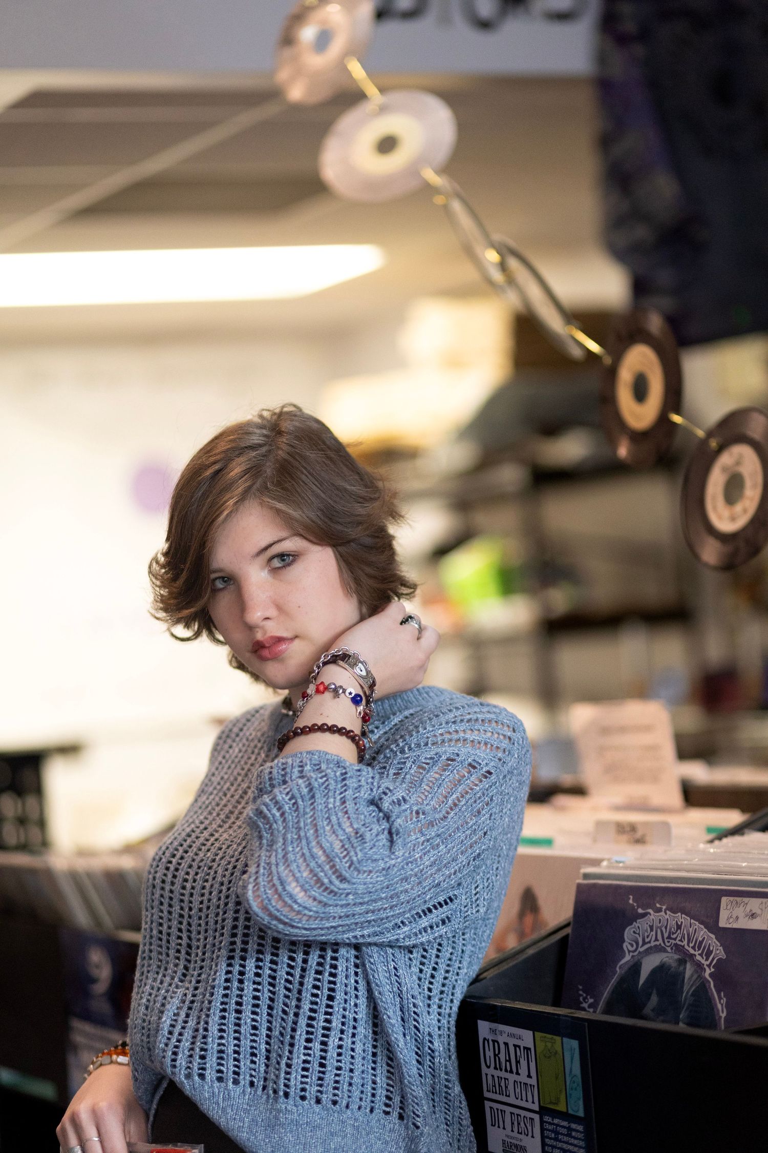Young woman in gray knit sweater posing in a record store with vinyl records displayed in background.
