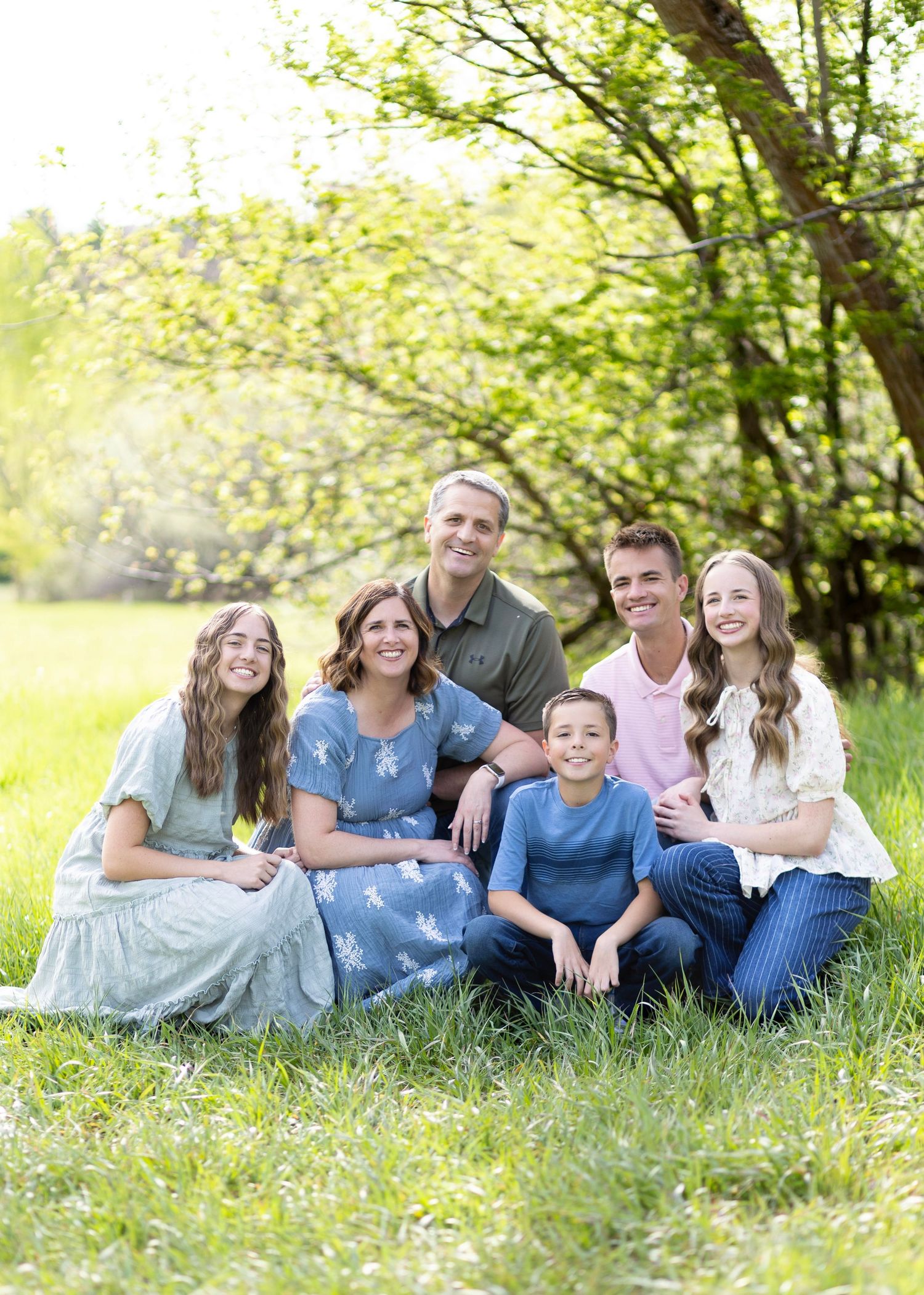 A happy family of six poses together on green grass under a blooming tree during a spring outdoor photo session.
