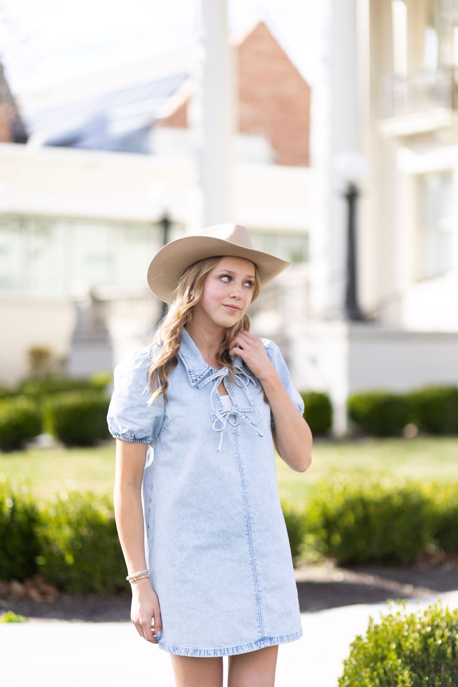 Young woman in light denim lace-up dress and tan wide-brim hat posing outdoors in urban setting.