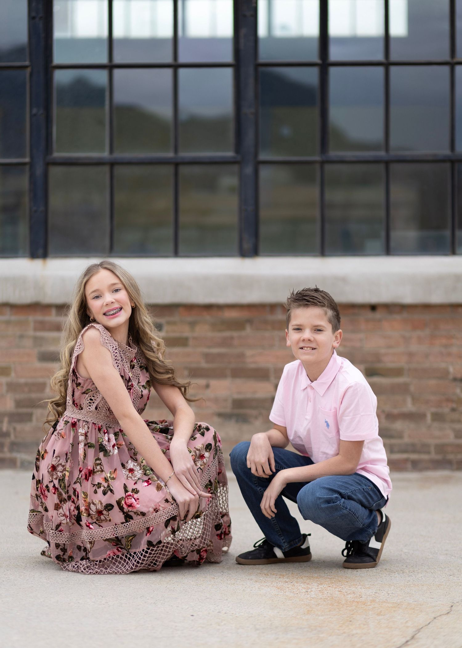 A girl in a floral dress and a boy in a pink polo shirt pose together outdoors near a brick wall.