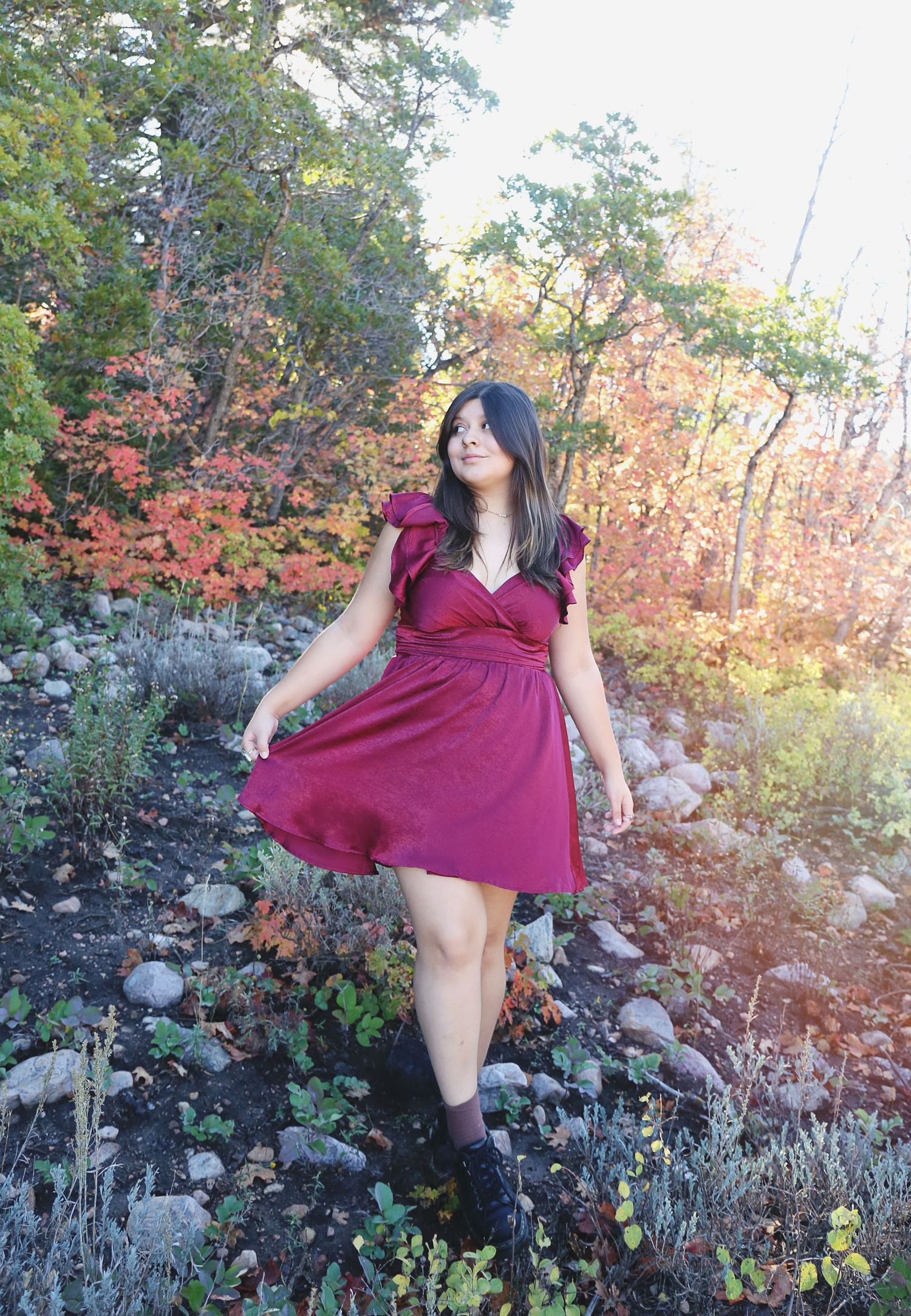 A person in a burgundy dress twirls in a garden setting with autumn foliage in the background.