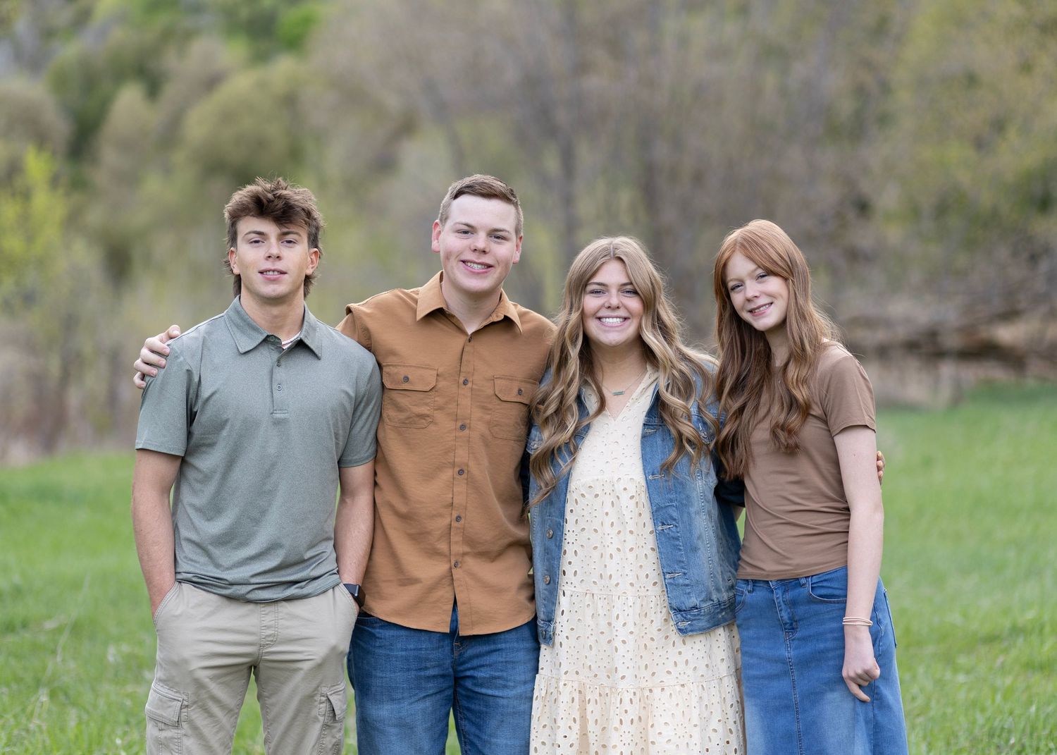 Four smiling young adults pose together in a green meadow for a casual outdoor family portrait.