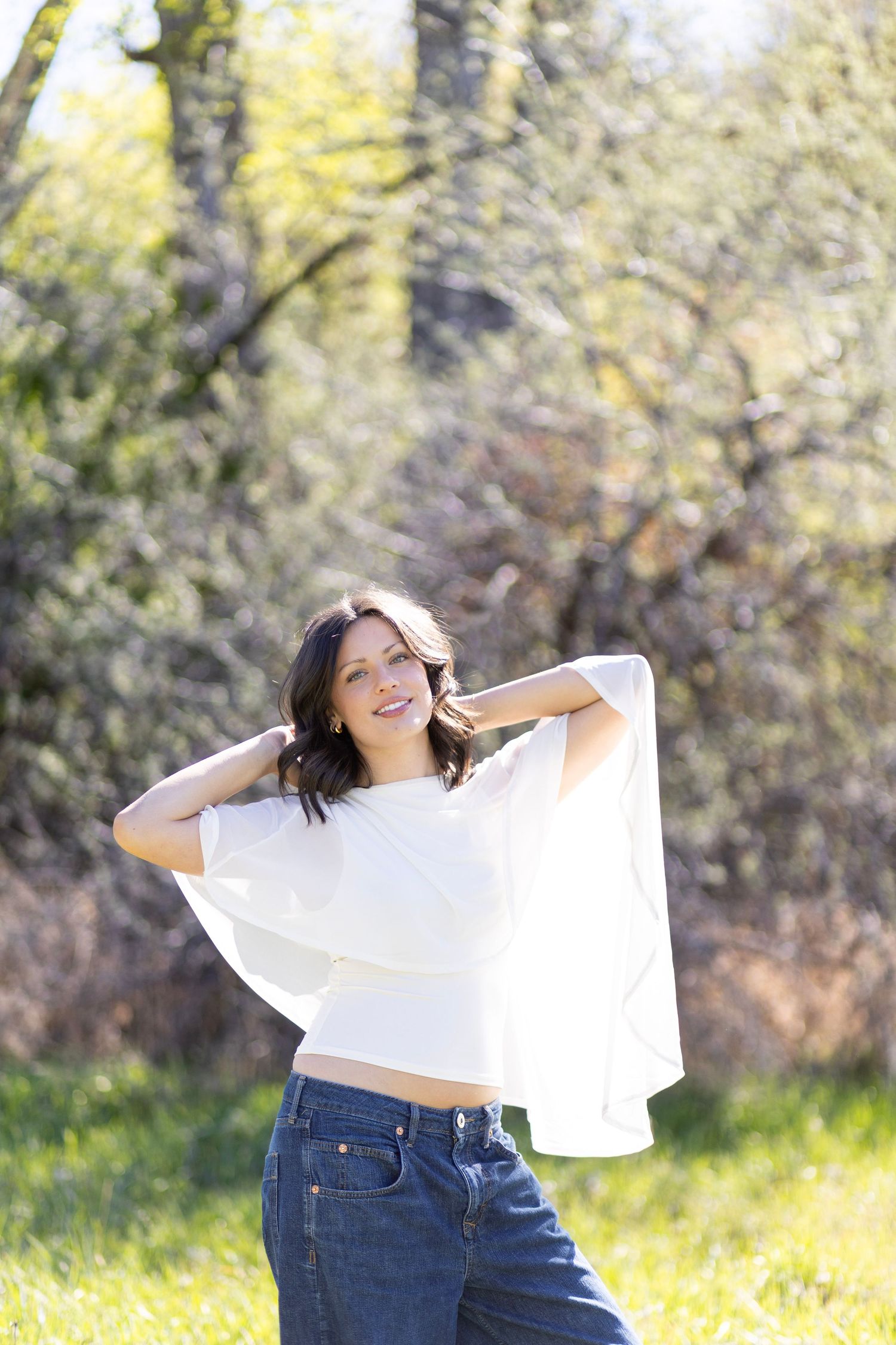 A smiling brunette woman in a white top and jeans poses outdoors in a sunny green park setting.