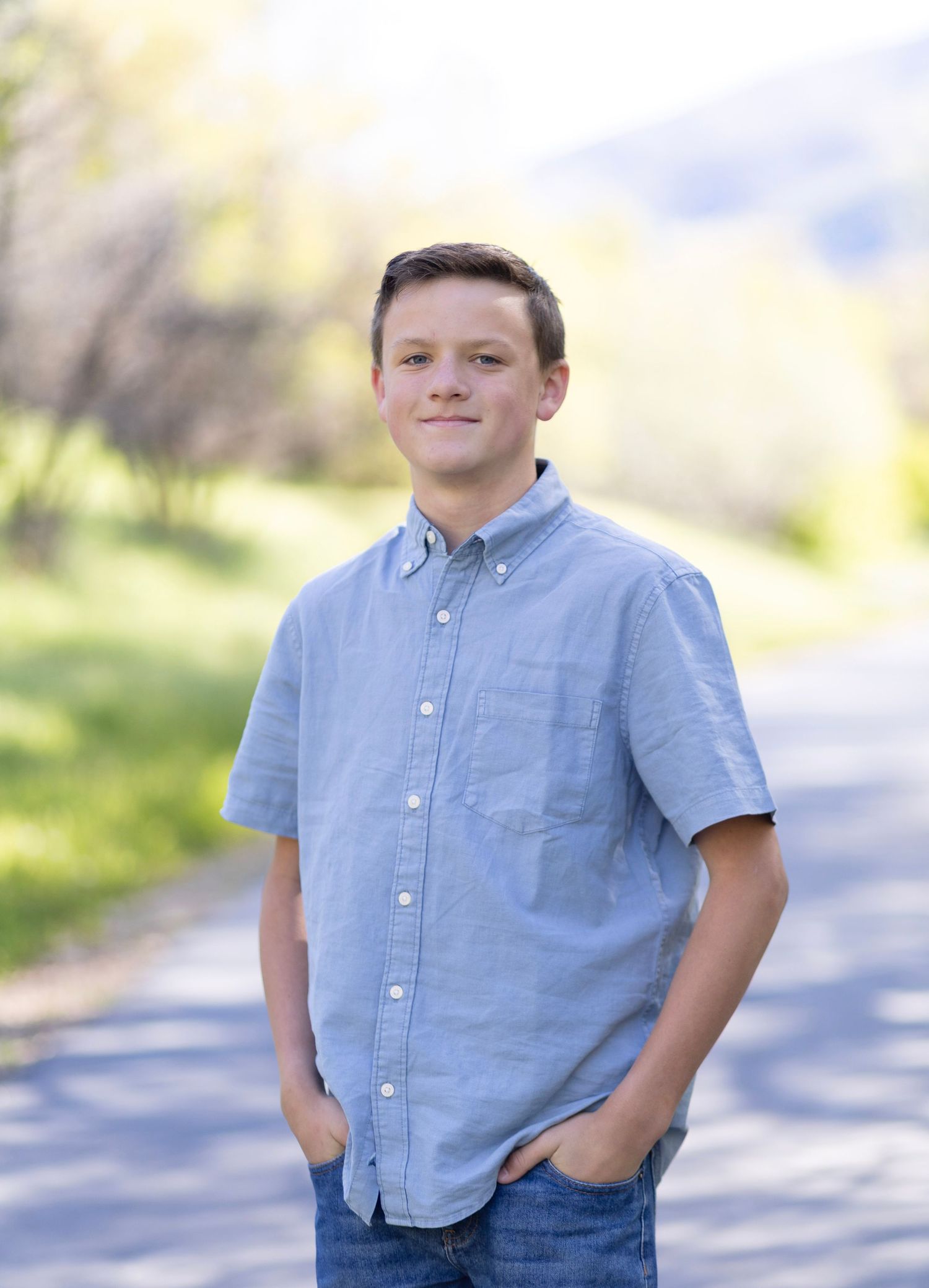 Smiling teenage boy in a light blue button-up shirt standing on a tree-lined path on a sunny day.