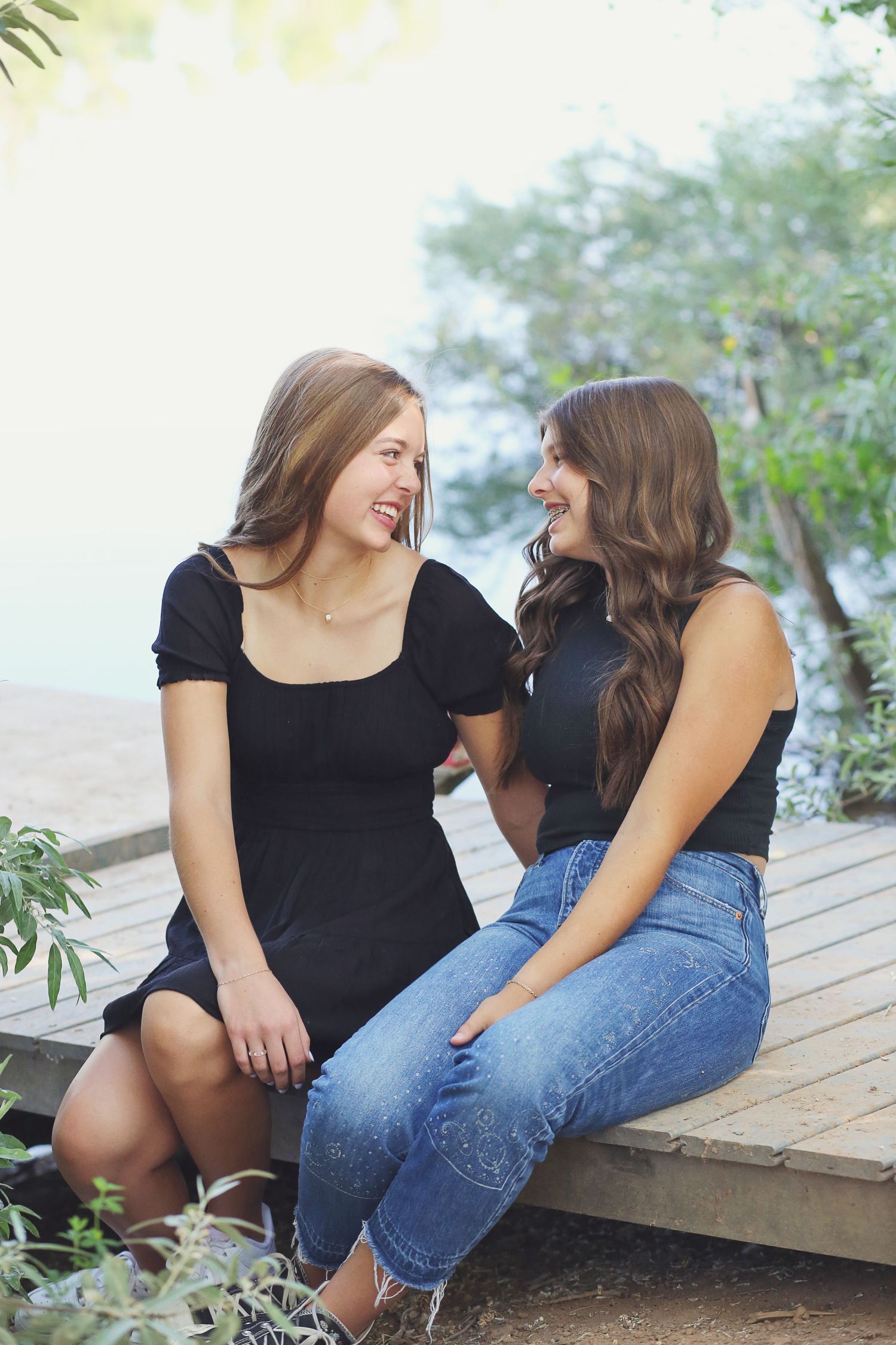 Two friends in casual outfits sitting and laughing together on wooden deck outdoors.