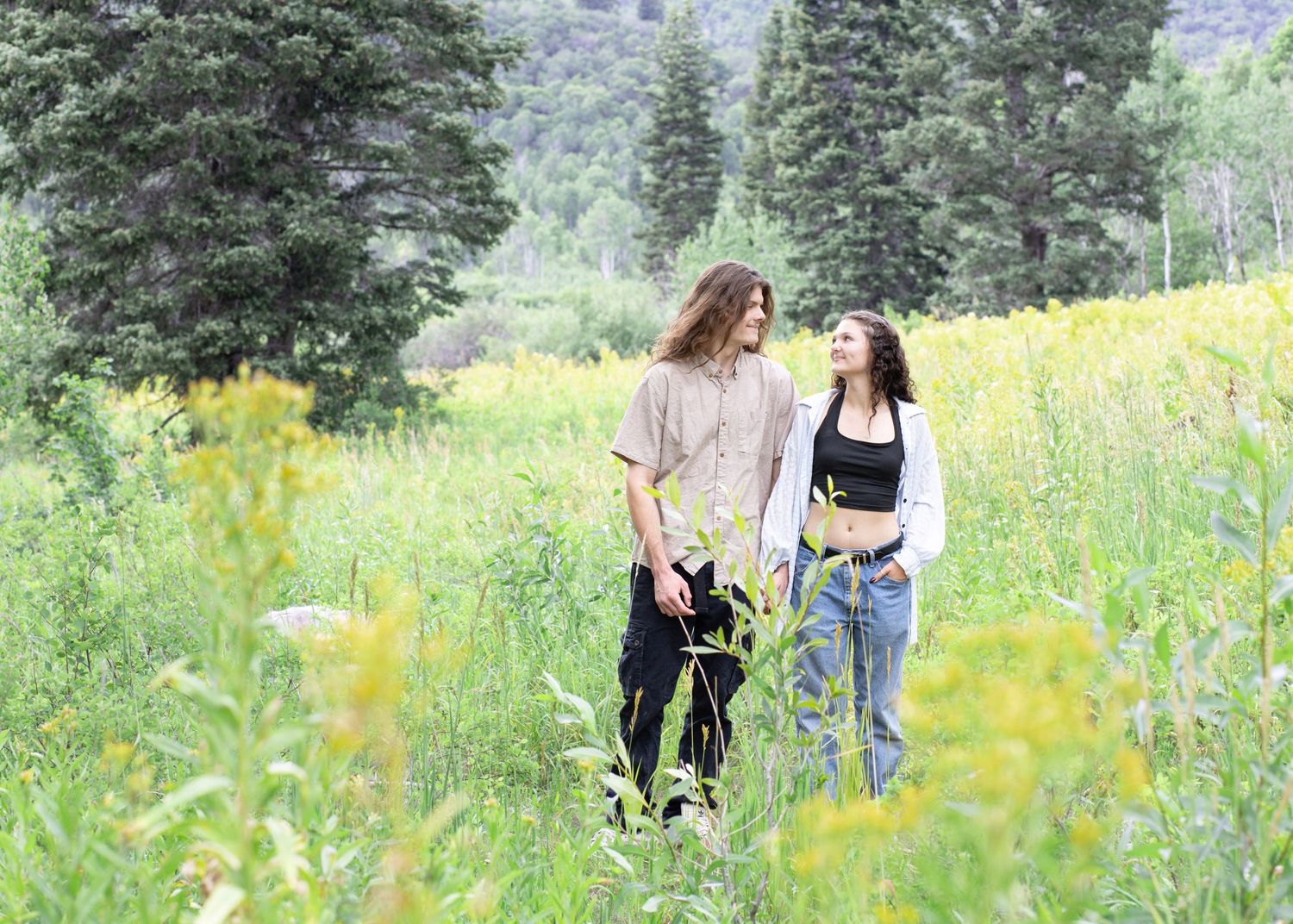 Couple walking through mountain wildflower field with evergreen forest backdrop.