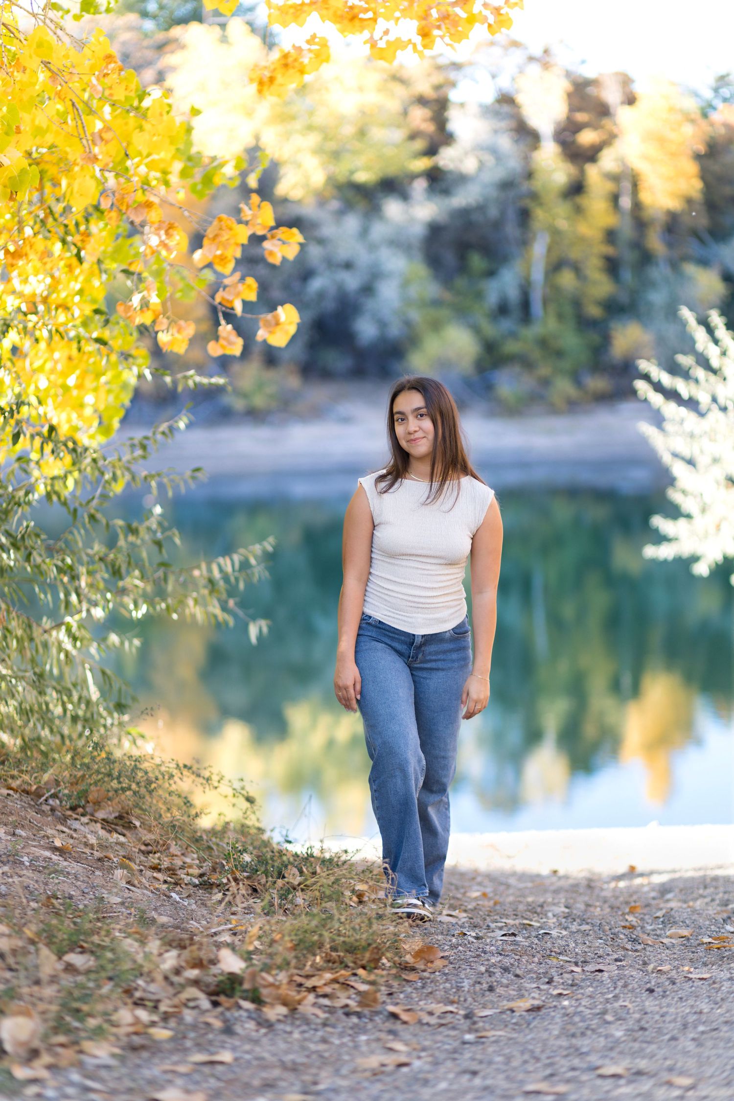 A person in casual attire sits by a serene lake surrounded by autumn trees with golden yellow foliage.