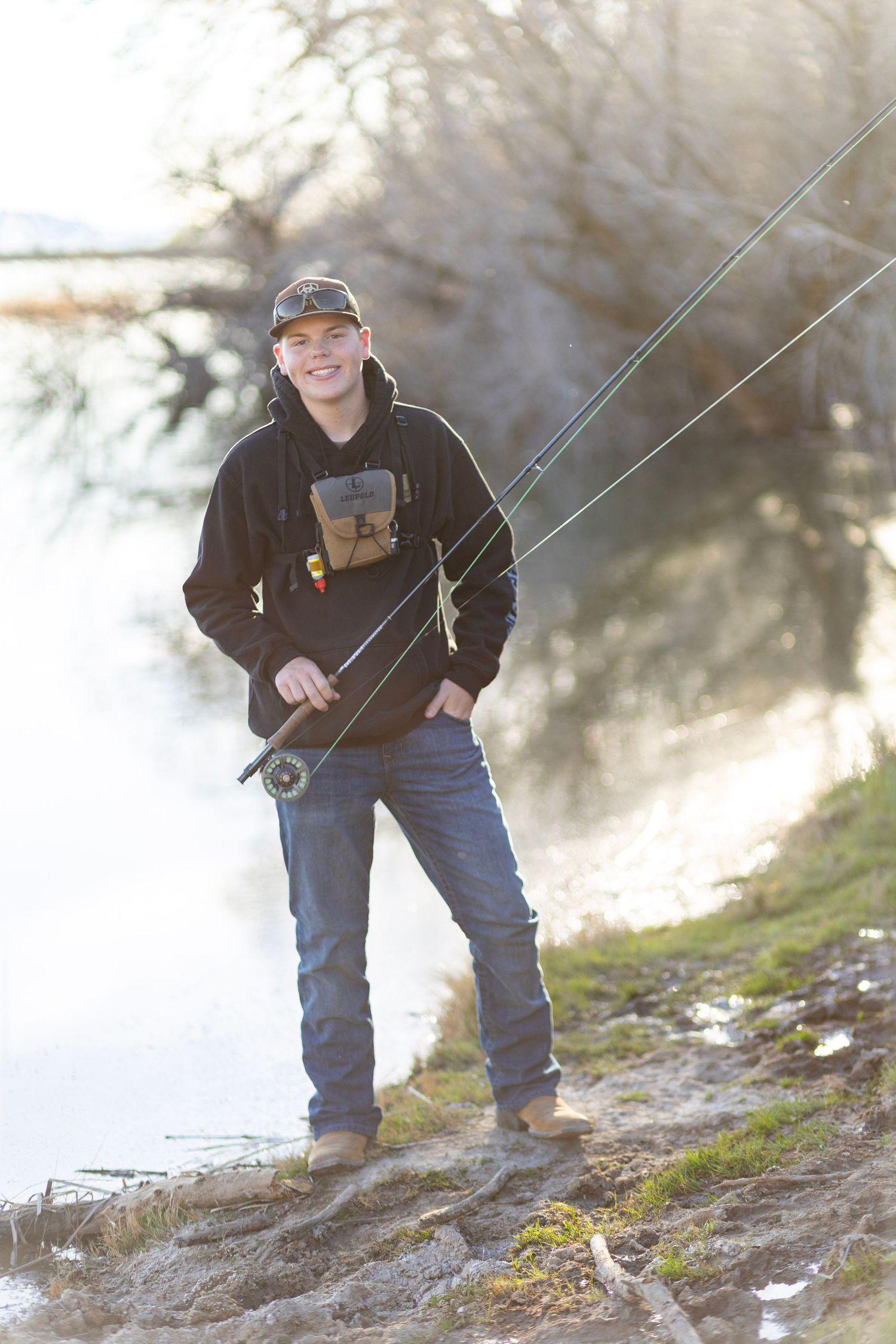 Smiling young man fly fishing by a misty river, wearing a black hoodie, cap, and jeans, holding a fishing rod.