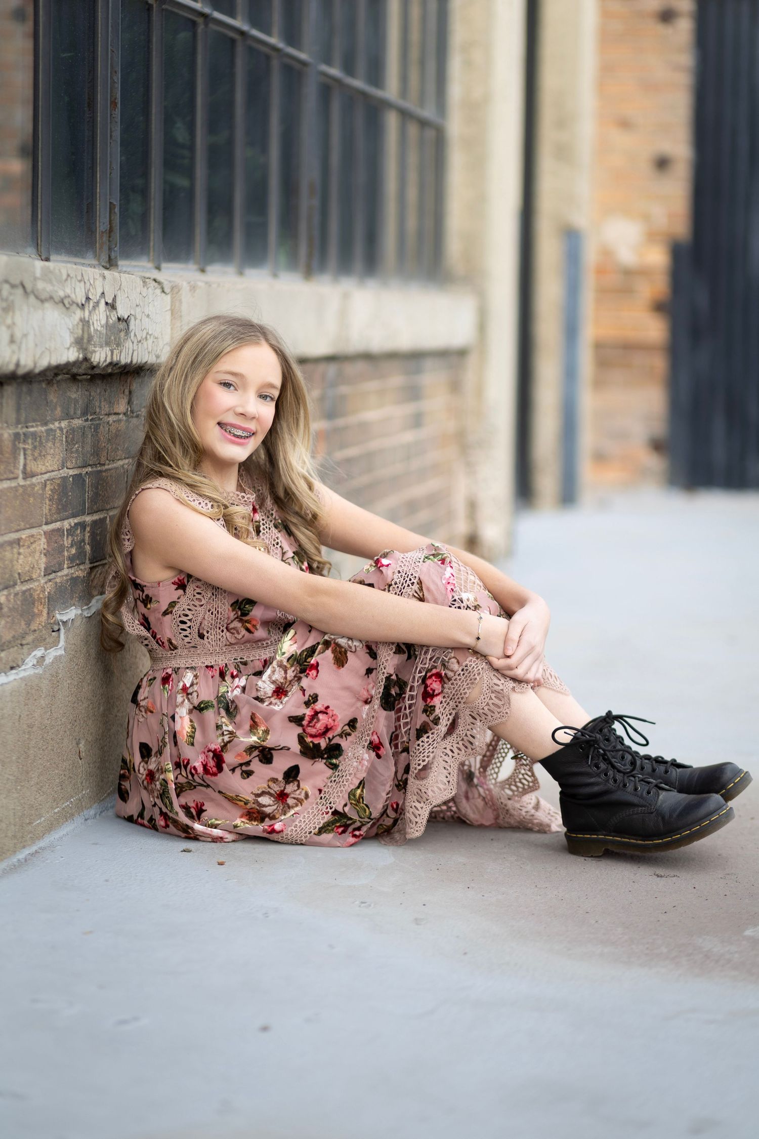 Young girl in floral dress and black boots sitting against brick wall, smiling outdoors.