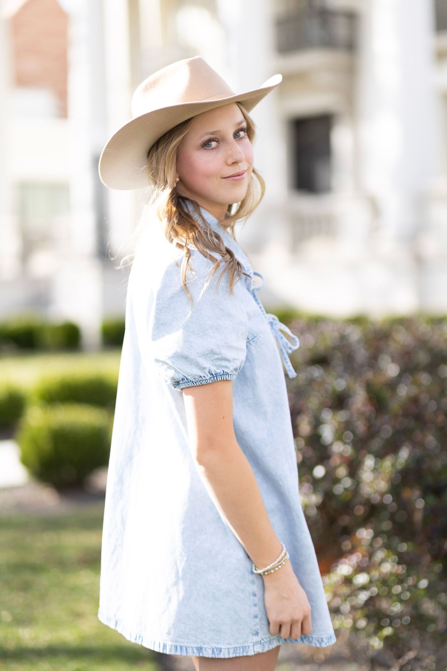 Young girl in light denim dress and tan cowboy hat poses outdoors near white building and green shrubs.