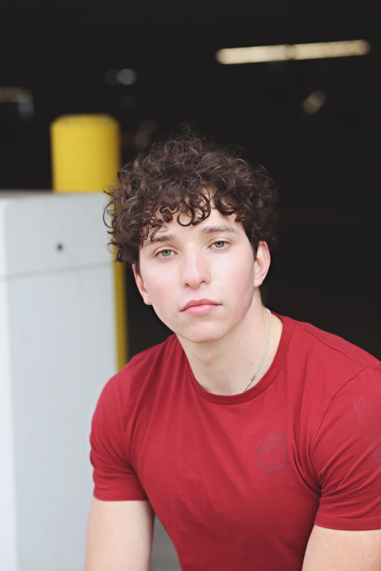 Portrait of a person with curly dark hair wearing a red t-shirt against a dark background.